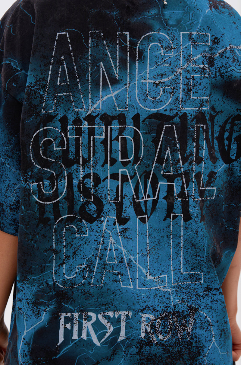 A person wears the ANCESTRAL FURY OVERSIZED GRAPHIC TEE in blue and black, showcasing its distressed, overlapping "ANGE SURBAN HISTORY CALL FIRST ROW" text on the back; only the shirt’s reverse and an arm are visible.