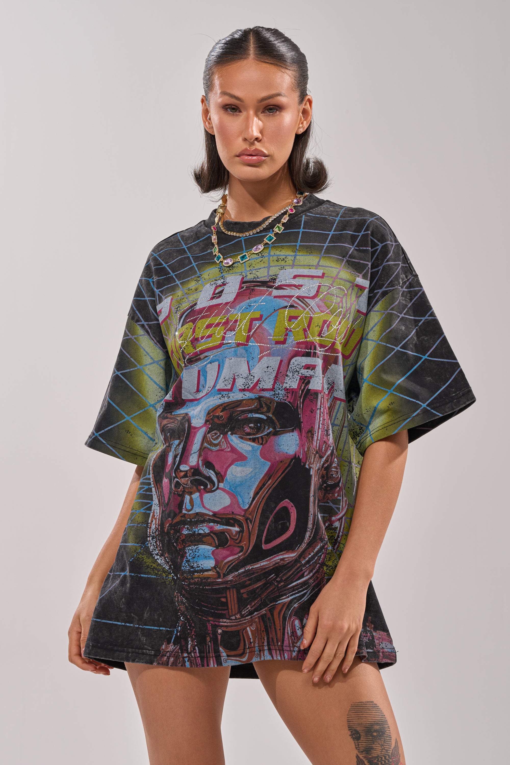 A woman with dark hair in pigtails wears the FIRST ROW NEW METAVERSE OVERSIZED GRAPHIC TEE featuring a bold, futuristic robot face and abstract patterns, embodying cyber style as she stands against a plain background. 0