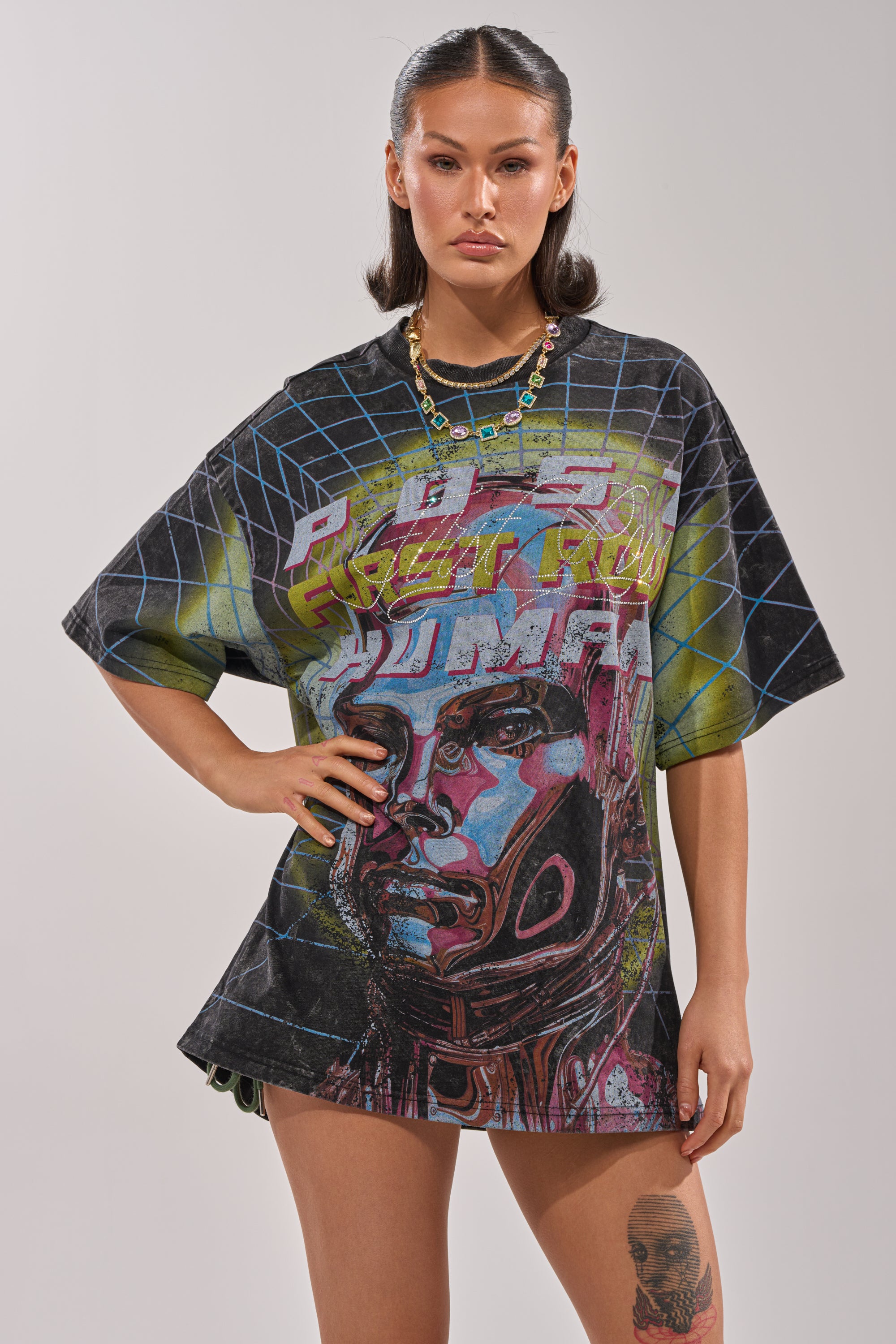 A woman exudes confidence in the FIRST ROW NEW METAVERSE OVERSIZED GRAPHIC TEE, featuring a futuristic colorful face and geometric patterns. She pairs it with slicked-back hair, a necklace, and showcases tattoos on her arm and thigh. 1