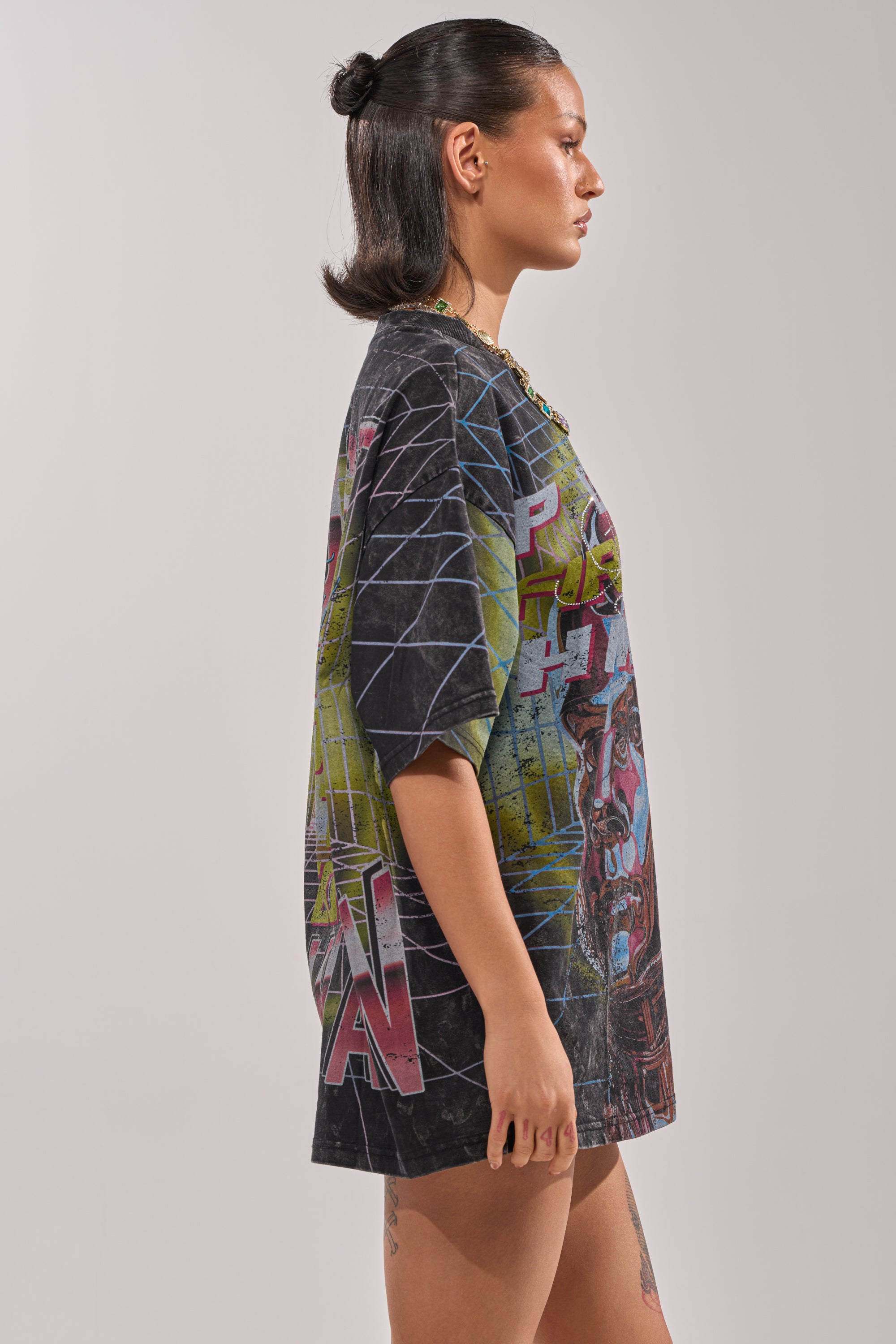 A woman with dark half-up, half-down hair stands in profile against a plain backdrop, wearing the FIRST ROW NEW METAVERSE OVERSIZED GRAPHIC TEE with colorful designs, styled with a bold chunky gold necklace for streetwear flair. 2
