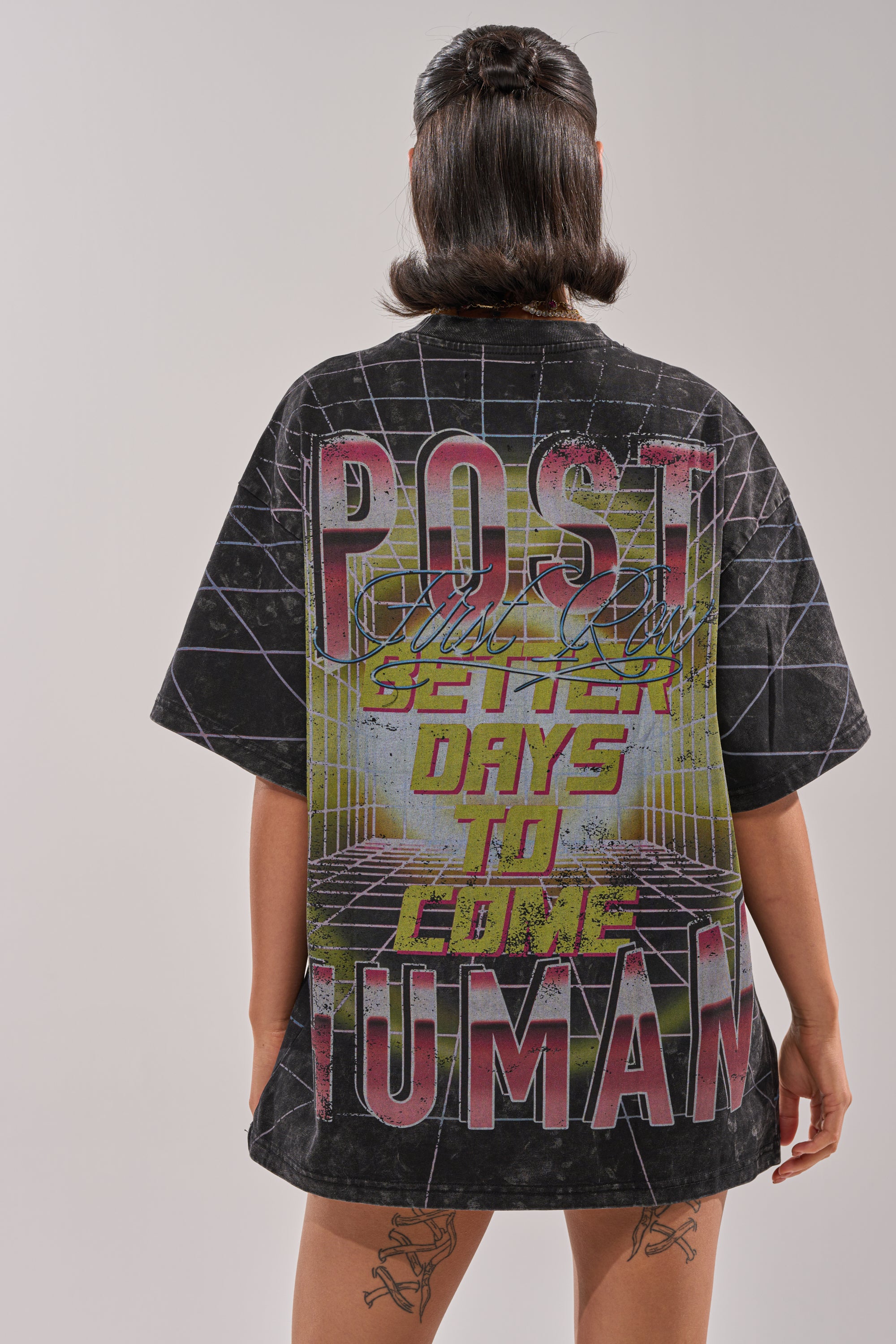 A person with short, dark hair wears the FIRST ROW NEW METAVERSE OVERSIZED GRAPHIC TEE featuring a retro-style "POST HUMAN SEVEN DAYS TO COME" print—ideal for cyber graphic streetwear fans. Tattoos are visible on their legs. 3