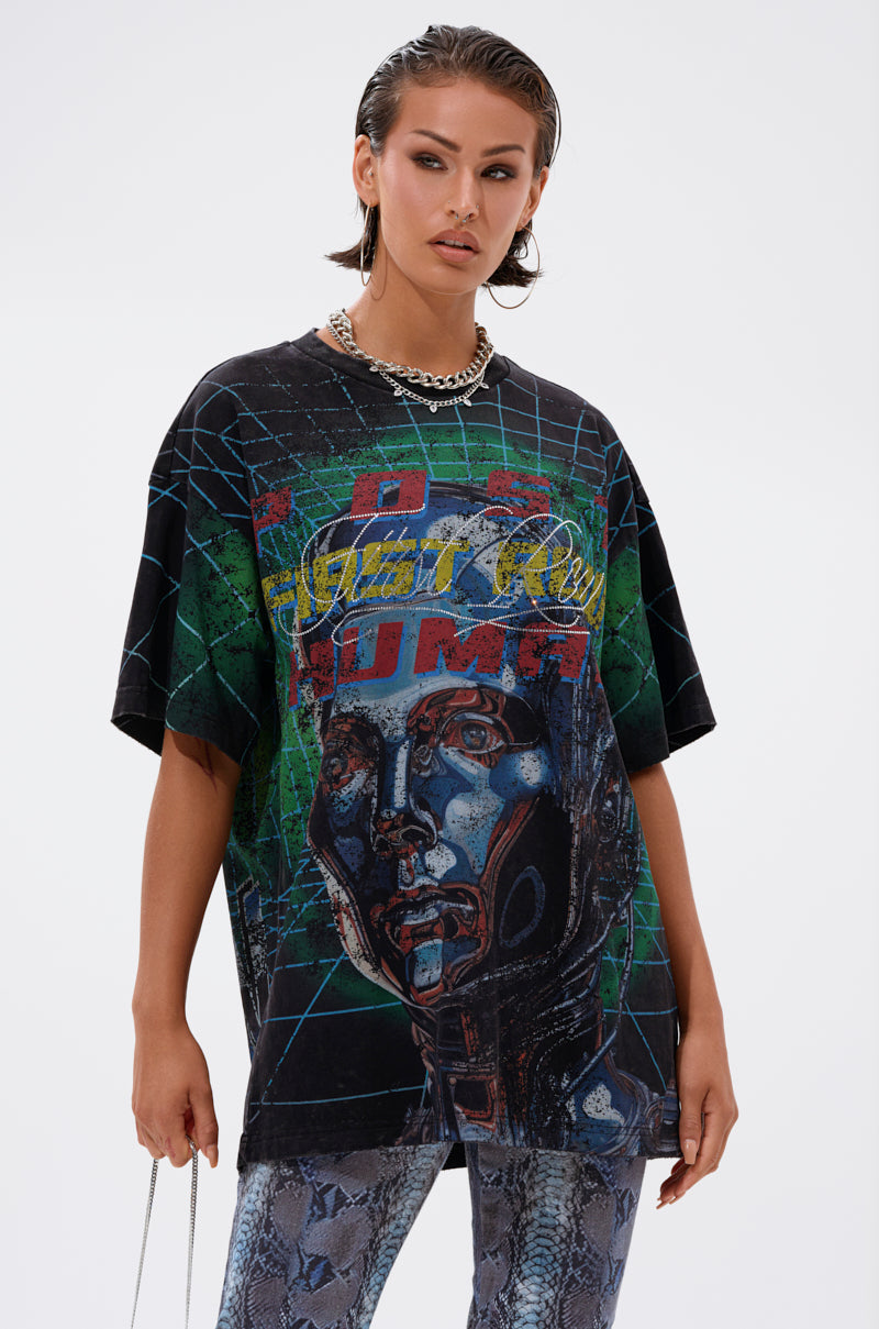 A woman with slicked-back hair wears the METAVERSE OVERSIZED GRAPHIC TEE, showcasing a futuristic robot-inspired graphic, paired with silver chain necklaces and patterned blue pants for a bold streetwear look against a plain backdrop.