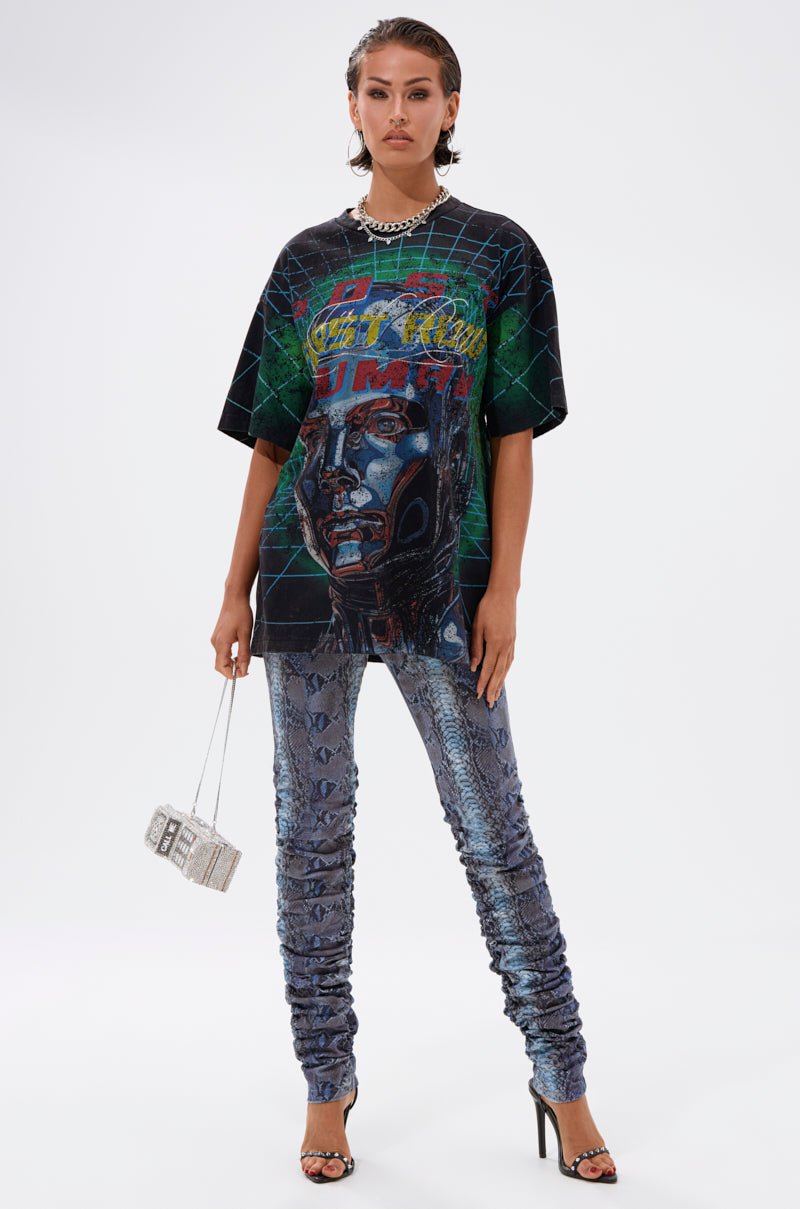 A woman confidently wears the METAVERSE OVERSIZED GRAPHIC TEE featuring a cyborg head, paired with shiny blue snakeskin pants, black heels, a silver purse, dramatic makeup, slicked-back hair, and a chunky necklace for bold streetwear flair.