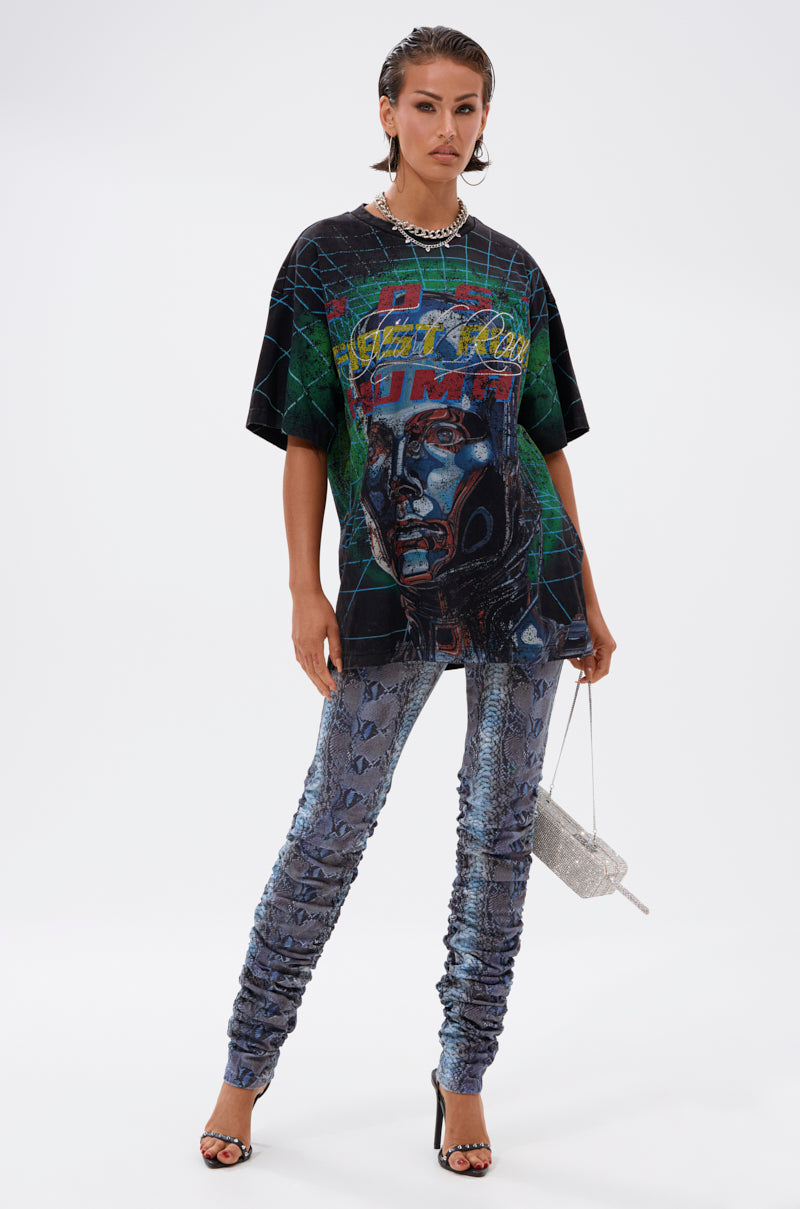 A woman poses against a plain white background wearing the METAVERSE OVERSIZED GRAPHIC TEE featuring a cyborg head graphic, styled with ruched snakeskin-print pants, black heels, a chunky chain necklace, and a sparkly silver handbag.