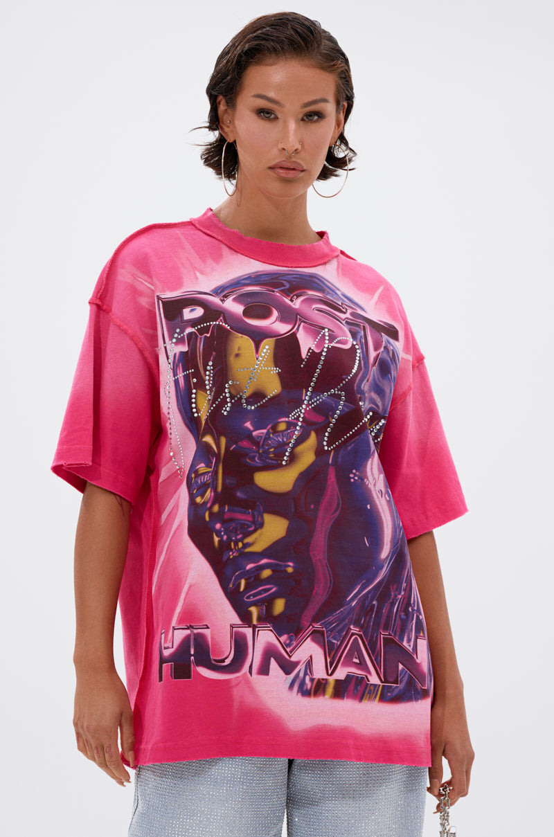 A woman with short wet-look hair wears the POST HUMAN OVERSIZED GRAPHIC TEE IN PINK, featuring a bold futuristic face graphic. She pairs it with light blue pants and hoop earrings, creating a striking streetwear look against a plain backdrop.