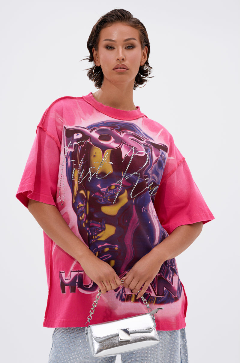 A person with short brown hair wears the POST HUMAN OVERSIZED GRAPHIC TEE IN PINK and holds a small, shiny silver handbag against a plain white background.