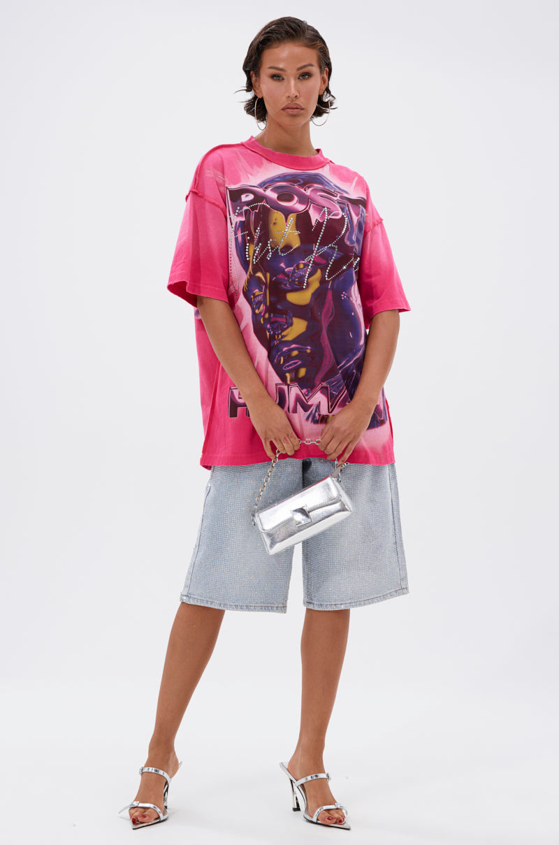 A woman stands against a white background wearing the POST HUMAN OVERSIZED GRAPHIC TEE IN PINK, light blue denim Bermuda shorts, silver high-heeled sandals, and holding a small metallic silver handbag.