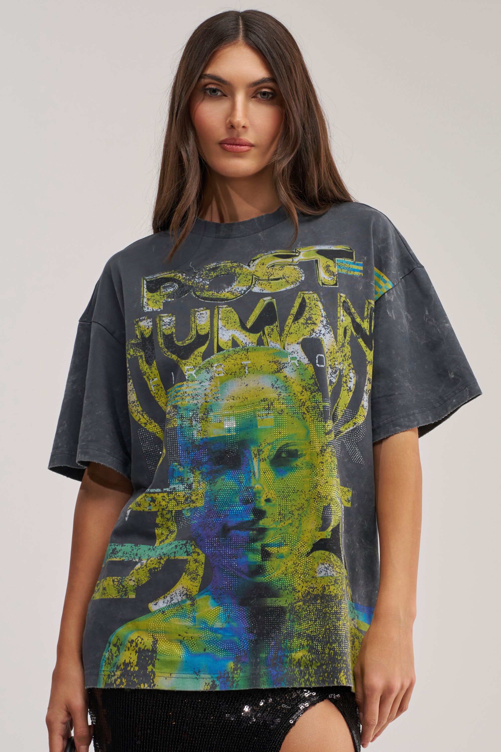 A woman with long brown hair wears the BETTER DAYS TO COME OVERSIZED GRAPHIC TEE, a washed grey cotton shirt featuring a colorful abstract face and "POST HUMAN" text. She stands facing forward with a neutral expression, ideal for summer streetwear.