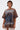 A person with short, wet-look hair wears the POST HUMAN OVERSIZED GRAPHIC TEE featuring blue and orange abstract art, paired with a white lace skirt—showcasing cyberpunk streetwear style against a plain light background.