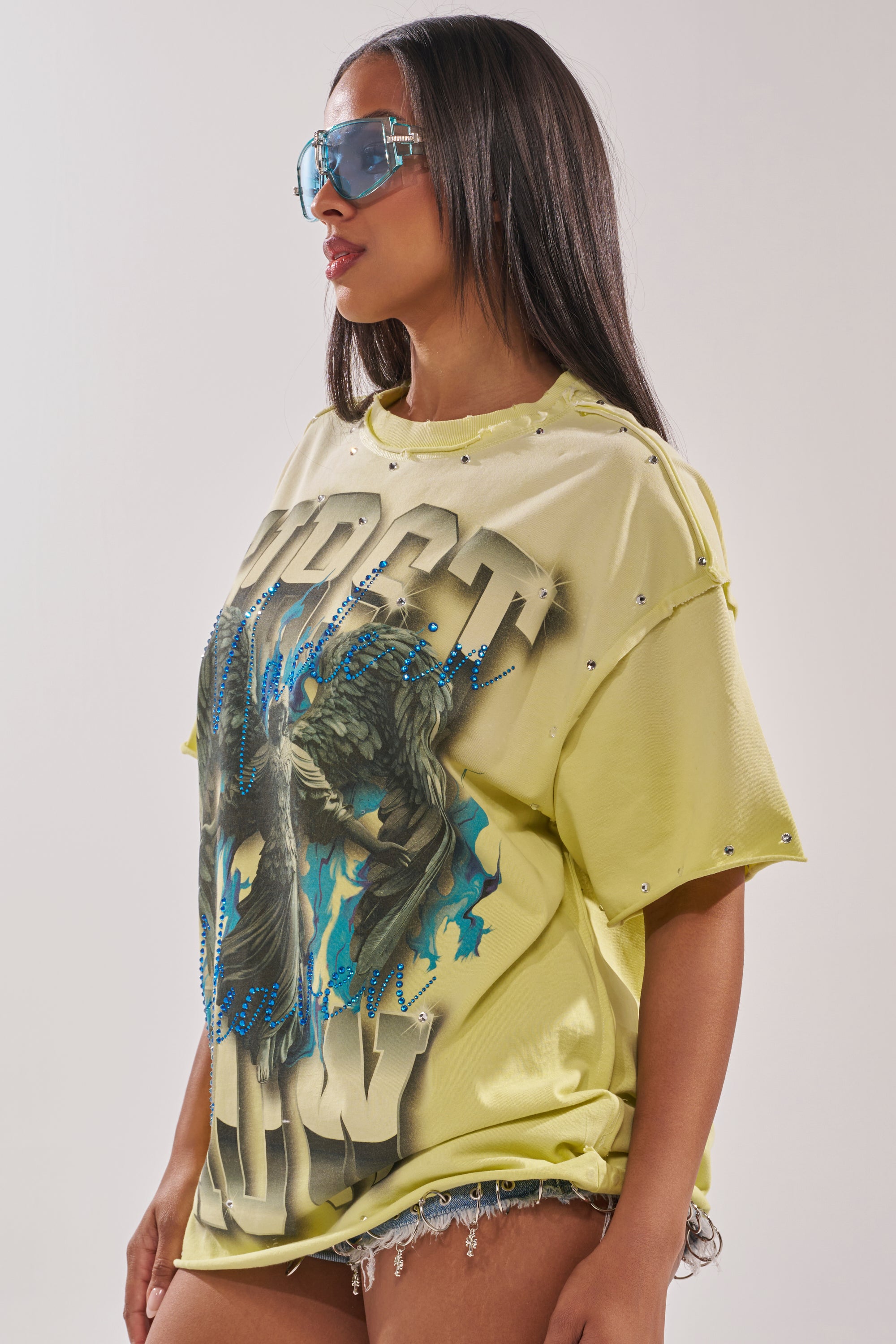 A woman with long straight hair wears oversized blue sunglasses, frayed denim shorts, and the FIRST ROW NEW RENAISSANCE RHINESTONE TEE—a loose yellow streetwear graphic T-shirt—standing against a plain light background. 2