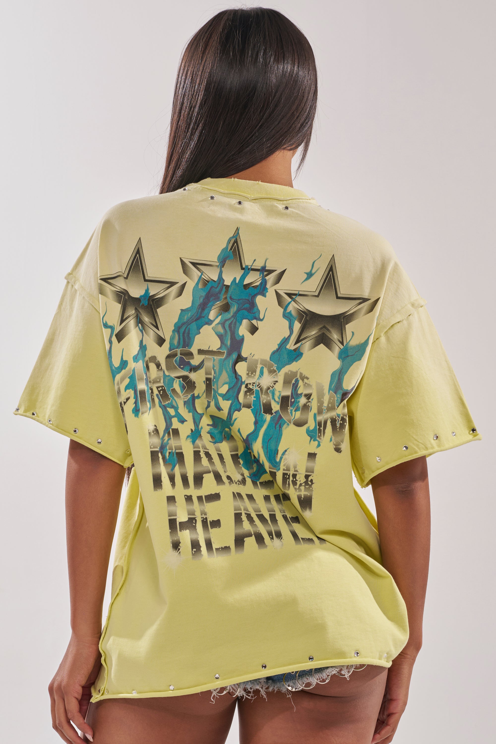 A person with long dark hair is seen from the back wearing the FIRST ROW NEW RENAISSANCE RHINESTONE TEE, a streetwear-inspired light yellow oversized shirt with star graphics, blue flames, bold black text, and denim shorts. 3