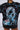 A person is wearing a RADICAL REVOLUTION TEE in black, crafted from soft cotton knit fabric and showcasing a digitally distorted image of a person's face with blue lightning bolts and red text. The head of the individual sporting the shirt is not visible in the photo.