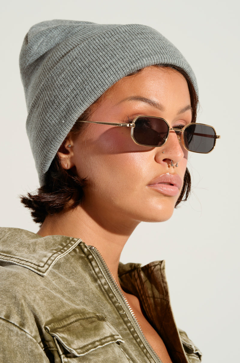 A person wearing a NEUTRAL GREY CLASSIC BEANIE for added warmth, along with sunglasses and an olive green jacket with the zipper partially open. The individual showcases a septum piercing and small hoop earrings, set against a plain, light-colored background.