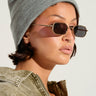 A person wearing a NEUTRAL GREY CLASSIC BEANIE for added warmth, along with sunglasses and an olive green jacket with the zipper partially open. The individual showcases a septum piercing and small hoop earrings, set against a plain, light-colored background.