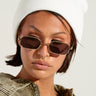 A person wearing a WINTER WHITE CLASSIC BEANIE, brown rectangular sunglasses, and a khaki jacket, with short dark hair and a septum piercing, looks downward against a light background.