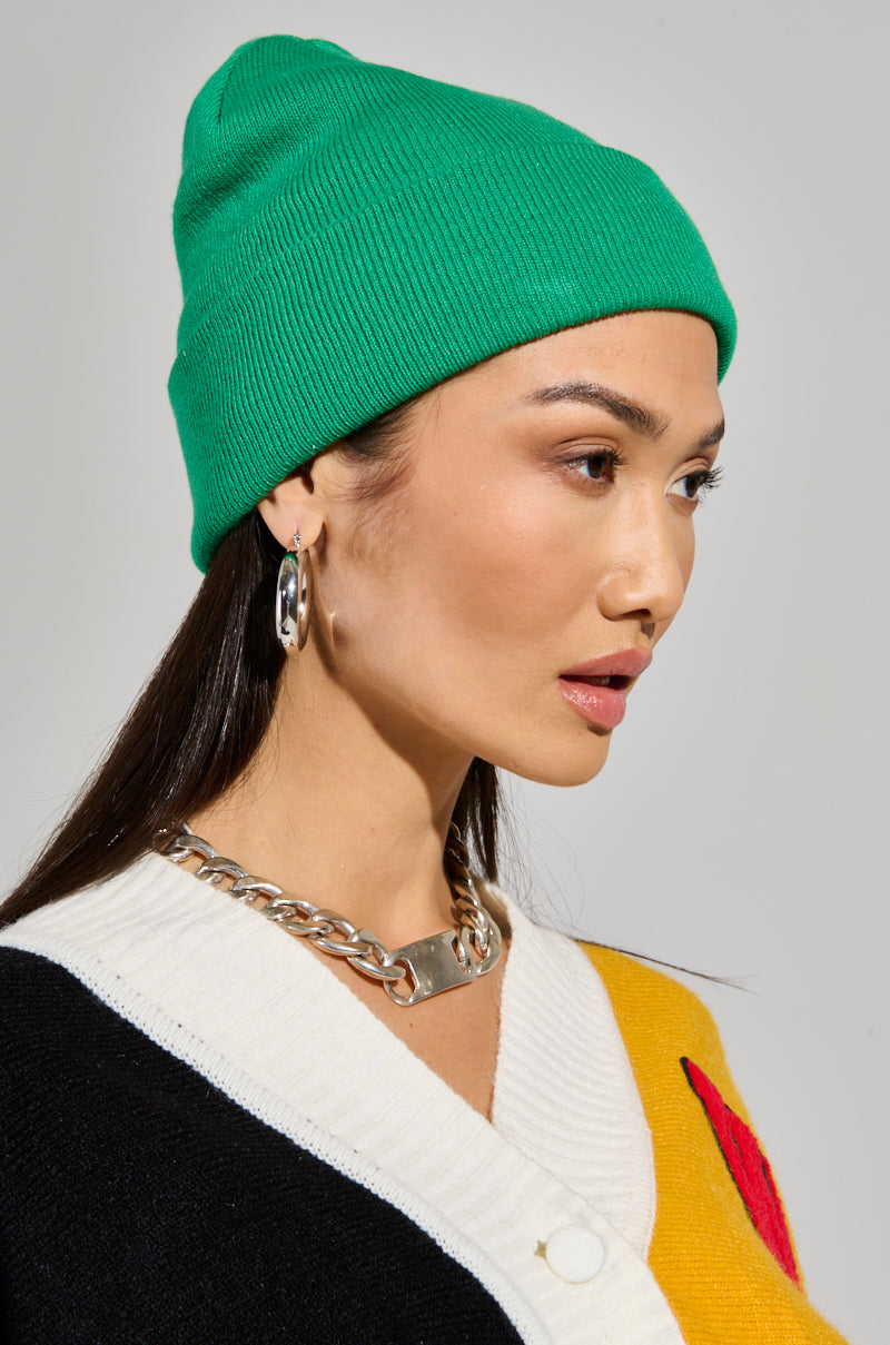 A person sporting the Classic Knit Beanie in green and silver hoop earrings looks to the side. They wear a color-blocked sweater featuring black, white, and mustard yellow sections. A chunky silver chain necklace completes the outfit.