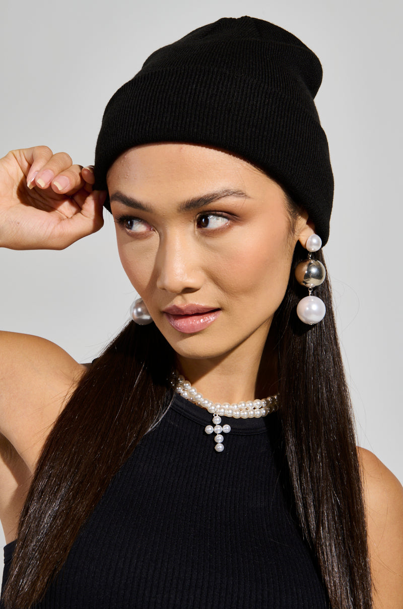 A person with long dark hair is wearing the BACK TO BLACK CLASSIC BEANIE and a black top, paired with pearl drop earrings and a pearl choker necklace. They are looking slightly to the side against a light gray backdrop.