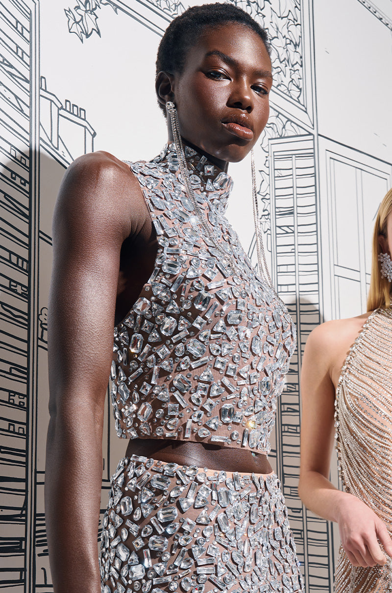 A model strikes a pose in the striking LOOKS SO GOOD TOP, featuring dazzling crystals and a sheer mesh design. In the background, there's an outline of a cityscape drawn in lines. Nearby, another individual is partially visible wearing an open-back design also embellished with rhinestones.