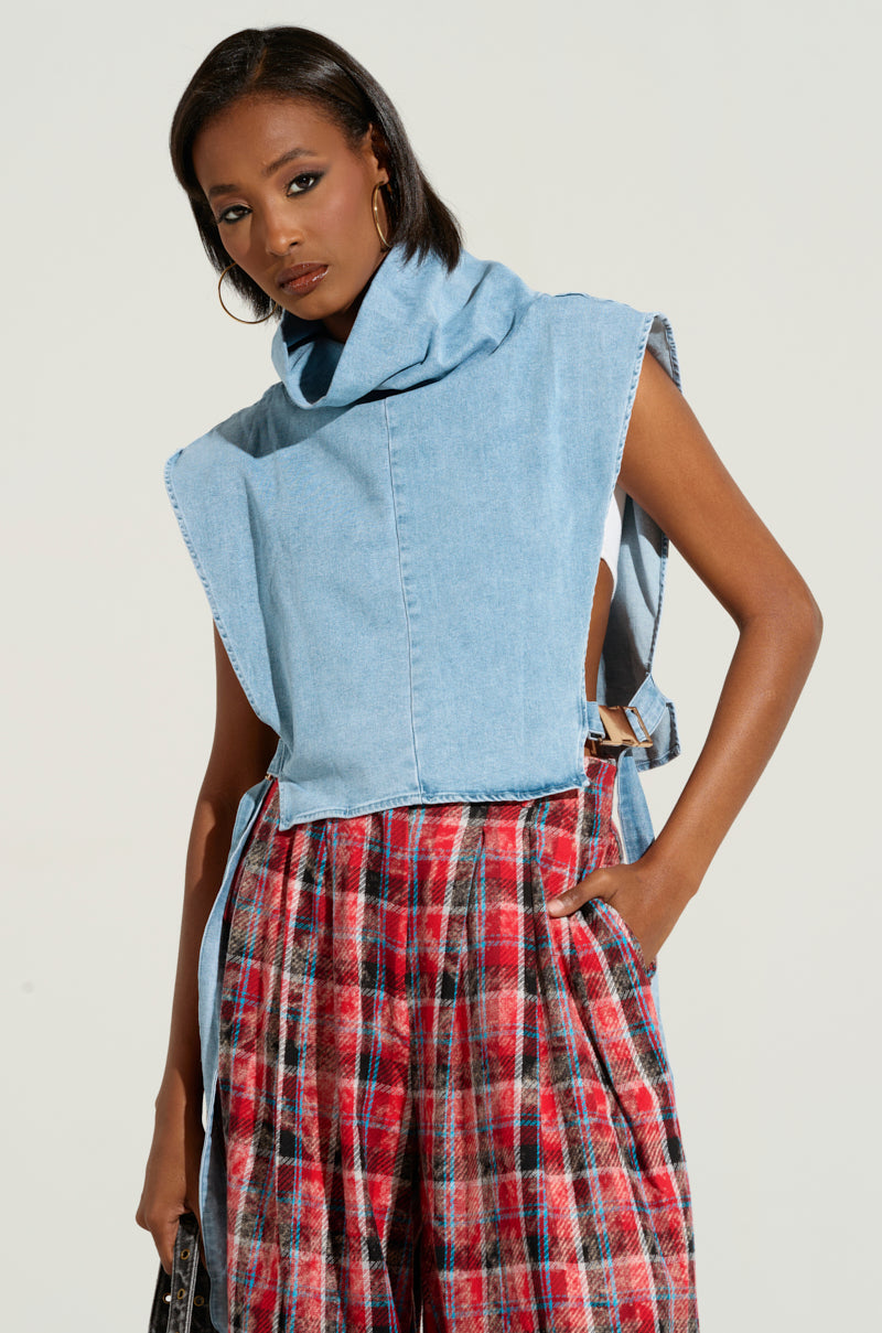 A person wearing the MY BEST SELF SLEEVELESS STATEMENT TOP in light blue denim with a high, wrap-around collar is also dressed in red and black plaid pants, with one hand in their pocket. The background is light grey and their expression is neutral, looking slightly to the side.