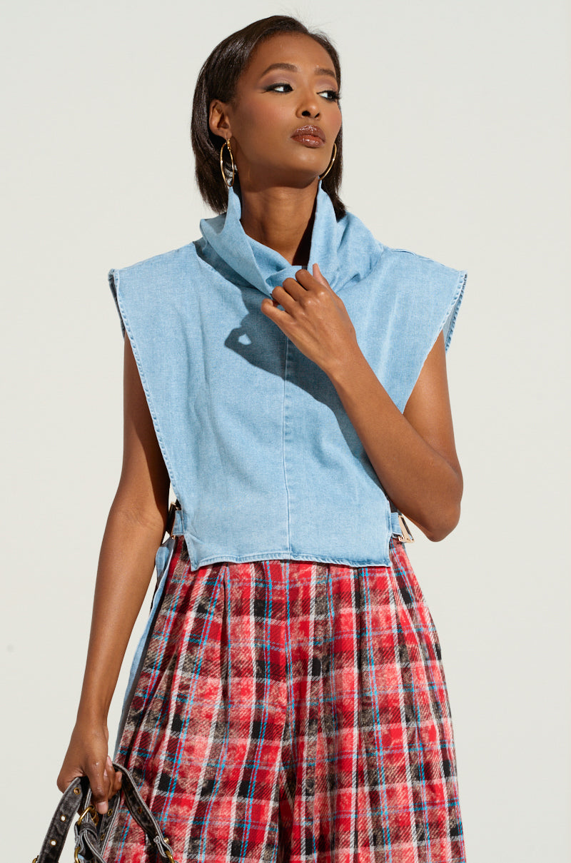 A person poses confidently against a plain background, wearing the MY BEST SELF SLEEVELESS STATEMENT TOP in light blue denim, featuring a high collar and adjustable side buckle closures. Paired with a red and blue plaid skirt, they have one hand near their chest and hold a handbag in the other hand. Hoop earrings complete the look.