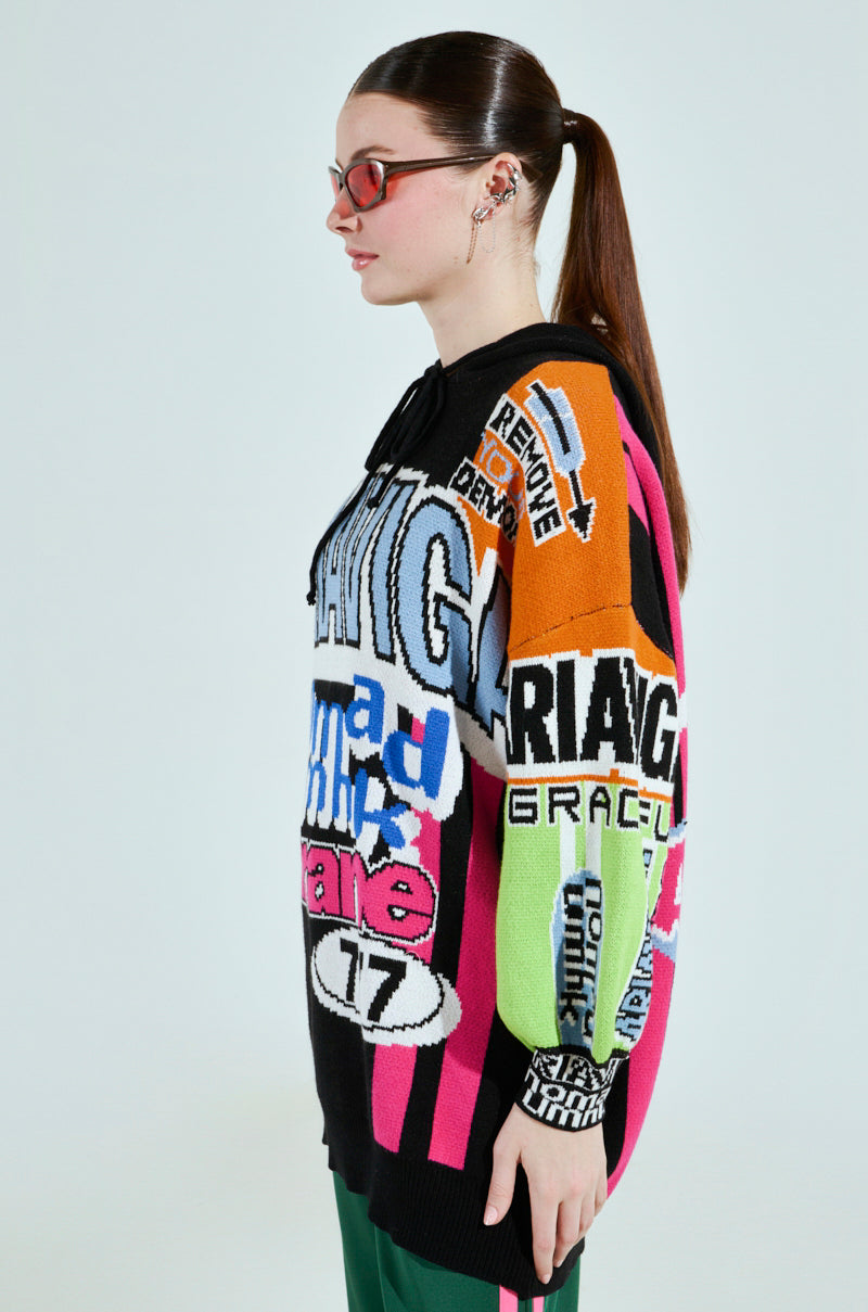 A person with long hair in a ponytail, red glasses, and an oversized JOYRIDE GRAPHIC HOODED SWEATER stands sideways against a plain background, showcasing its vibrant colorblocking and eye-catching mix of text and patterns.