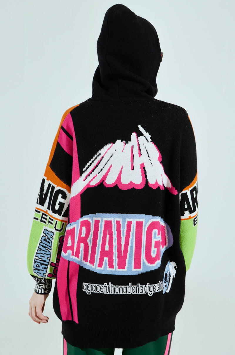 Back view of someone wearing the JOYRIDE GRAPHIC HOODED SWEATER, an oversized hoodie with vibrant pink, black, orange, and green colorblocking. It features abstract designs and bold text like "ARIAVIG" along with patterned elements.