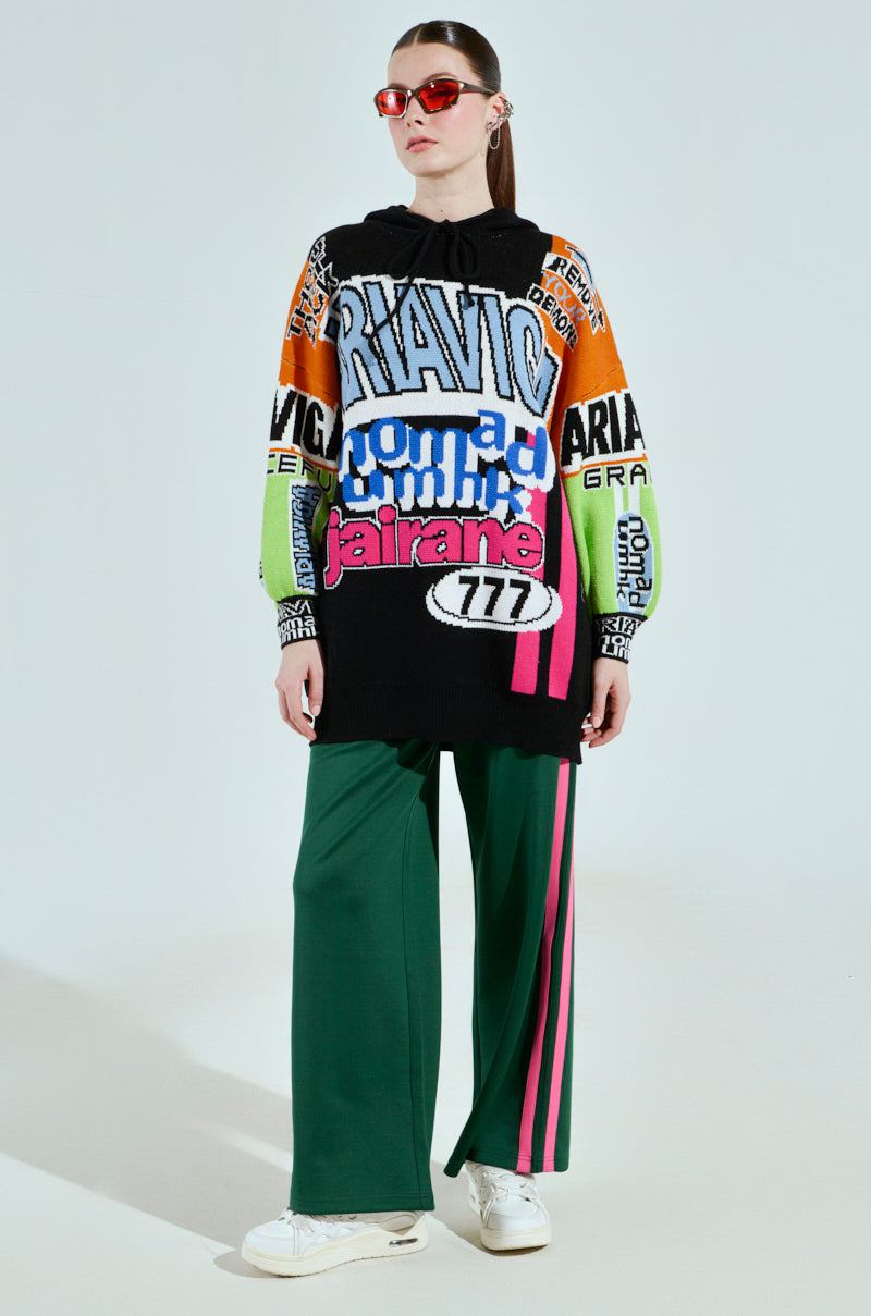 Wearing the JOYRIDE GRAPHIC HOODED SWEATER with vibrant colorblocking, paired with green wide-leg pants featuring pink stripes, red sunglasses, and white sneakers complete the look against a light background.