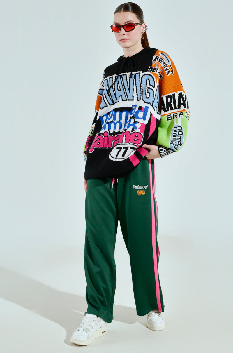 A person wears the vibrant JOYRIDE GRAPHIC HOODED SWEATER paired with green track pants featuring pink stripes, white sneakers, and red sunglasses, standing against a light background.