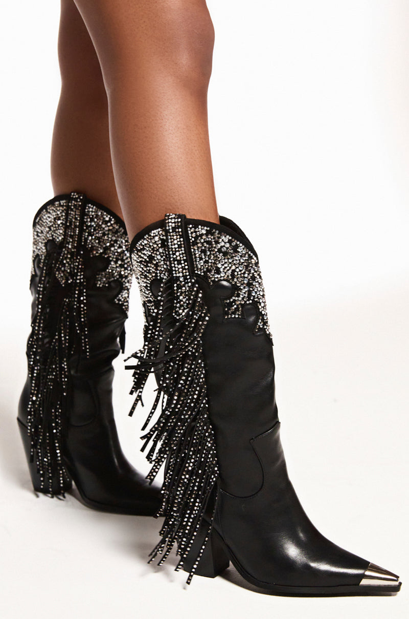 A close-up shows someone wearing the AZALEA WANG FULLER black rhinestone embellished cowboy boots, complete with pointed toes, shiny fringe details, metallic accents, and a chunky heel. These faux leather boots add sparkle against a plain white background.