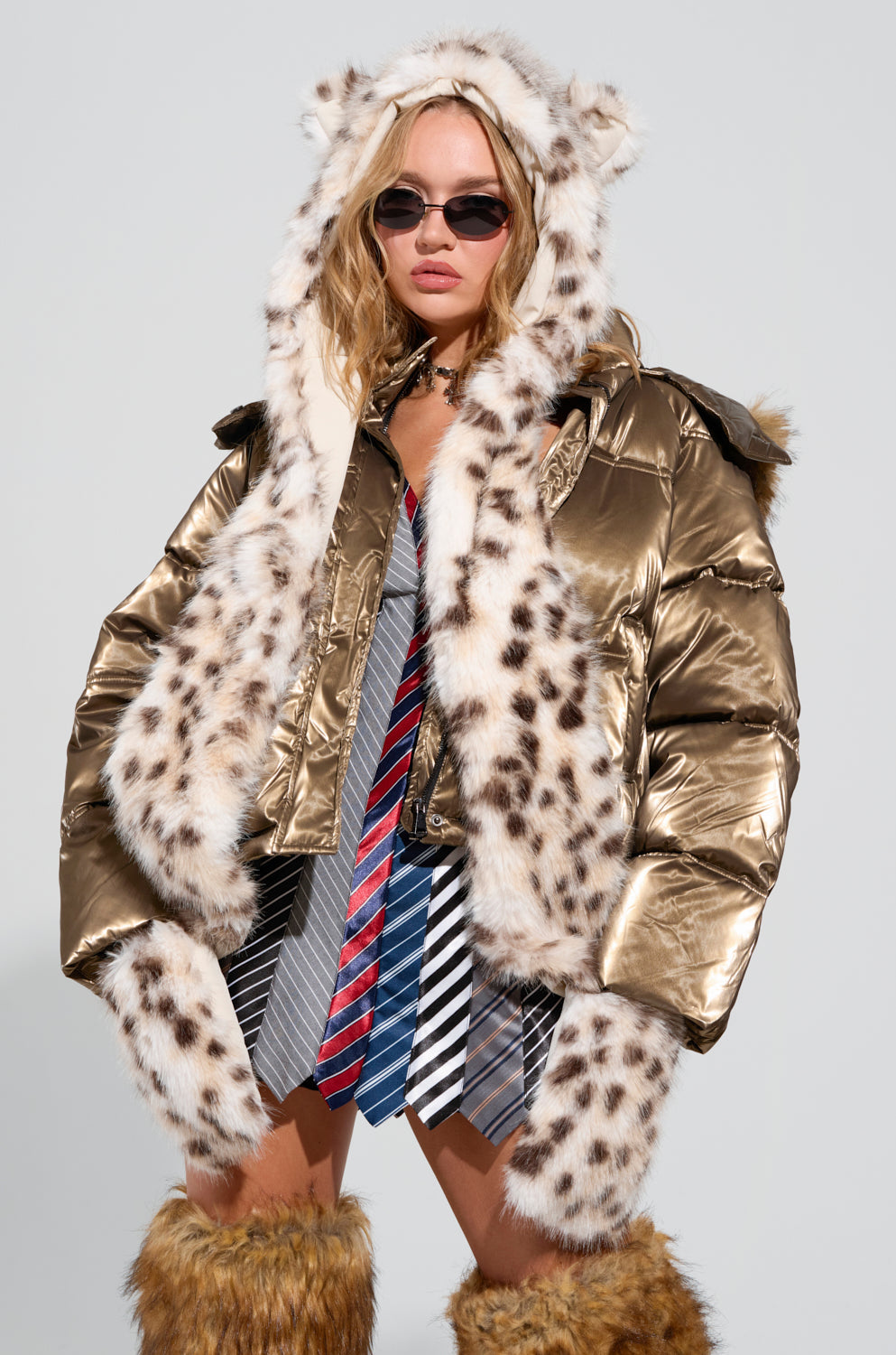A person stands confidently in the ANIME DREAMIN FAUX FUR HOOD, a gold puffer jacket with a kawaii faux fur hood featuring snow leopard print trim. They sport sunglasses and furry boots, complemented by a layered skirt made of colorful neckties against a simple, light gray backdrop. 0