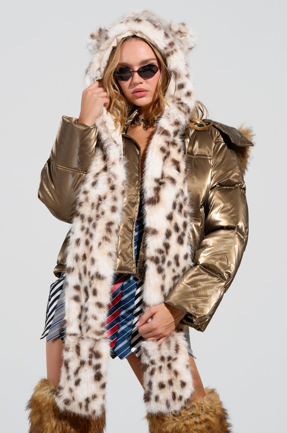 A person wearing the ANIME DREAMIN FAUX FUR HOOD, a metallic gold puffer jacket with a snow leopard print hood and matching long scarf. They accessorize with sunglasses, a plaid skirt, and fur-trimmed boots against a plain backdrop. 1