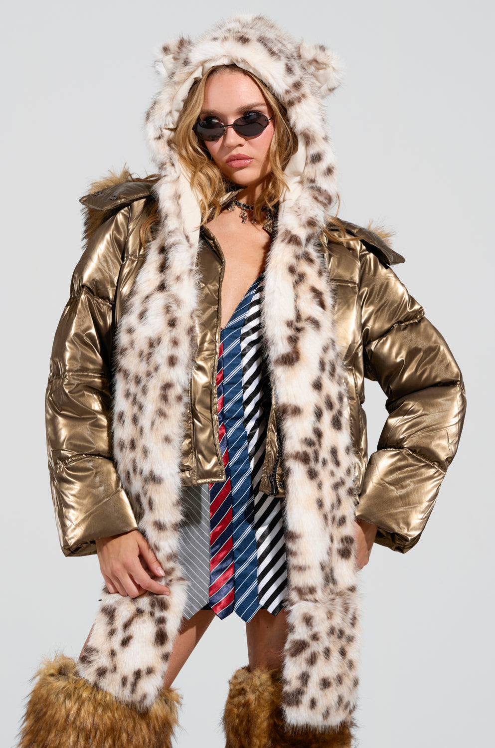 A person is sporting an Anime Dreamin Faux Fur Hood in gold with a snow leopard print, paired with sunglasses and a blue striped shirt featuring tie accents. The look is completed with fur-trimmed boots against a plain background. 2