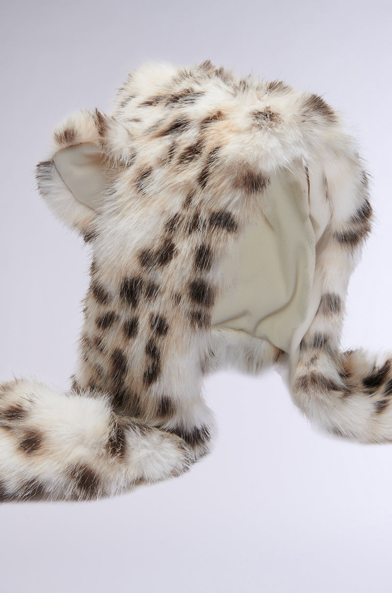 The ANIME DREAMIN FAUX FUR HOOD features a kawaii snow leopard print with ear flaps and a soft cream inner lining. Its long, dangling flaps contribute to a wild, cozy aesthetic against a simple backdrop. 9