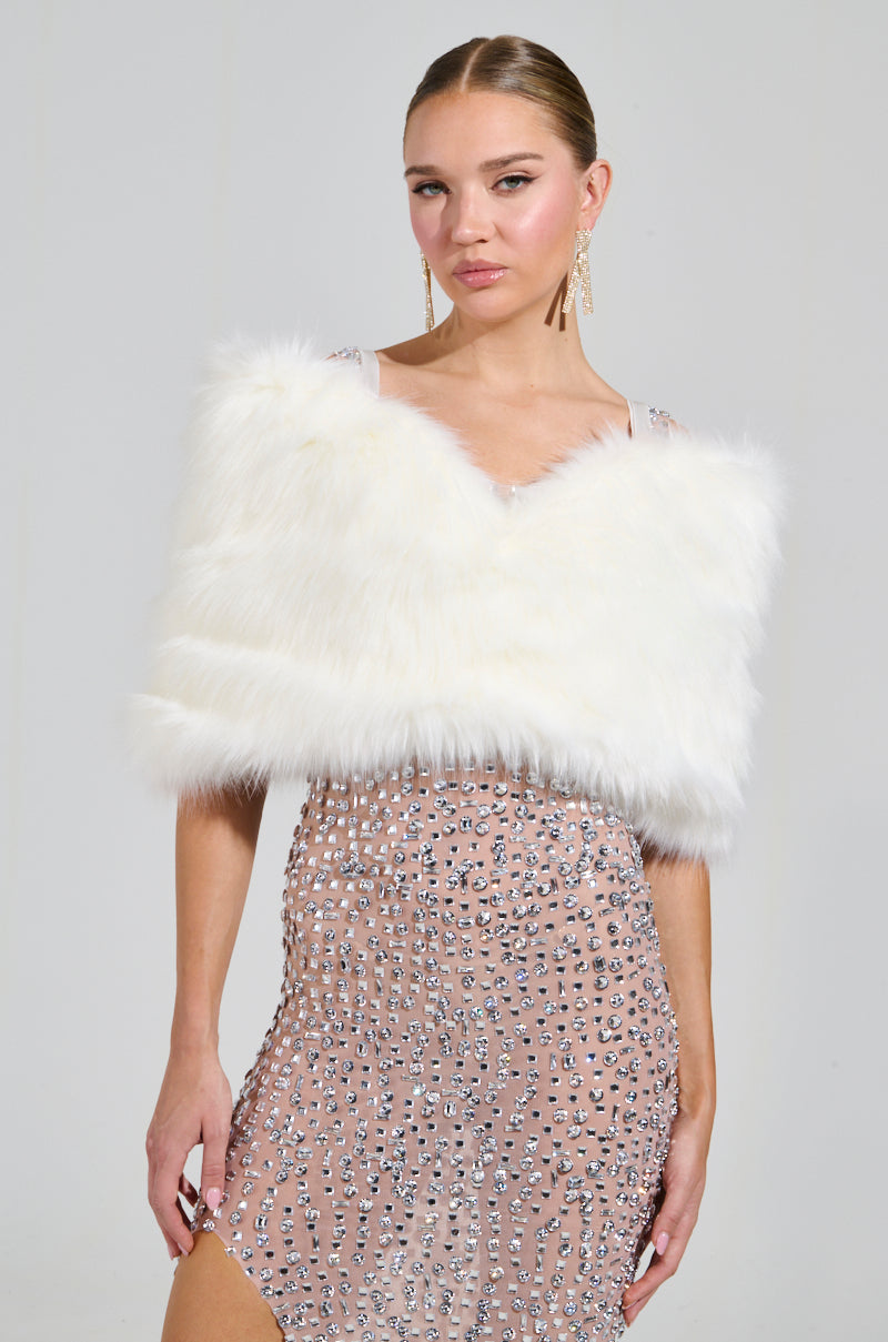 A woman wearing a sleeveless, embellished nude dress adorned with sparkling details pairs it with the elegant TO THE GALA FAUX FUR SHAWL off-the-shoulder. Her long earrings enhance her slicked-back hair as she poses against a simple backdrop. 2