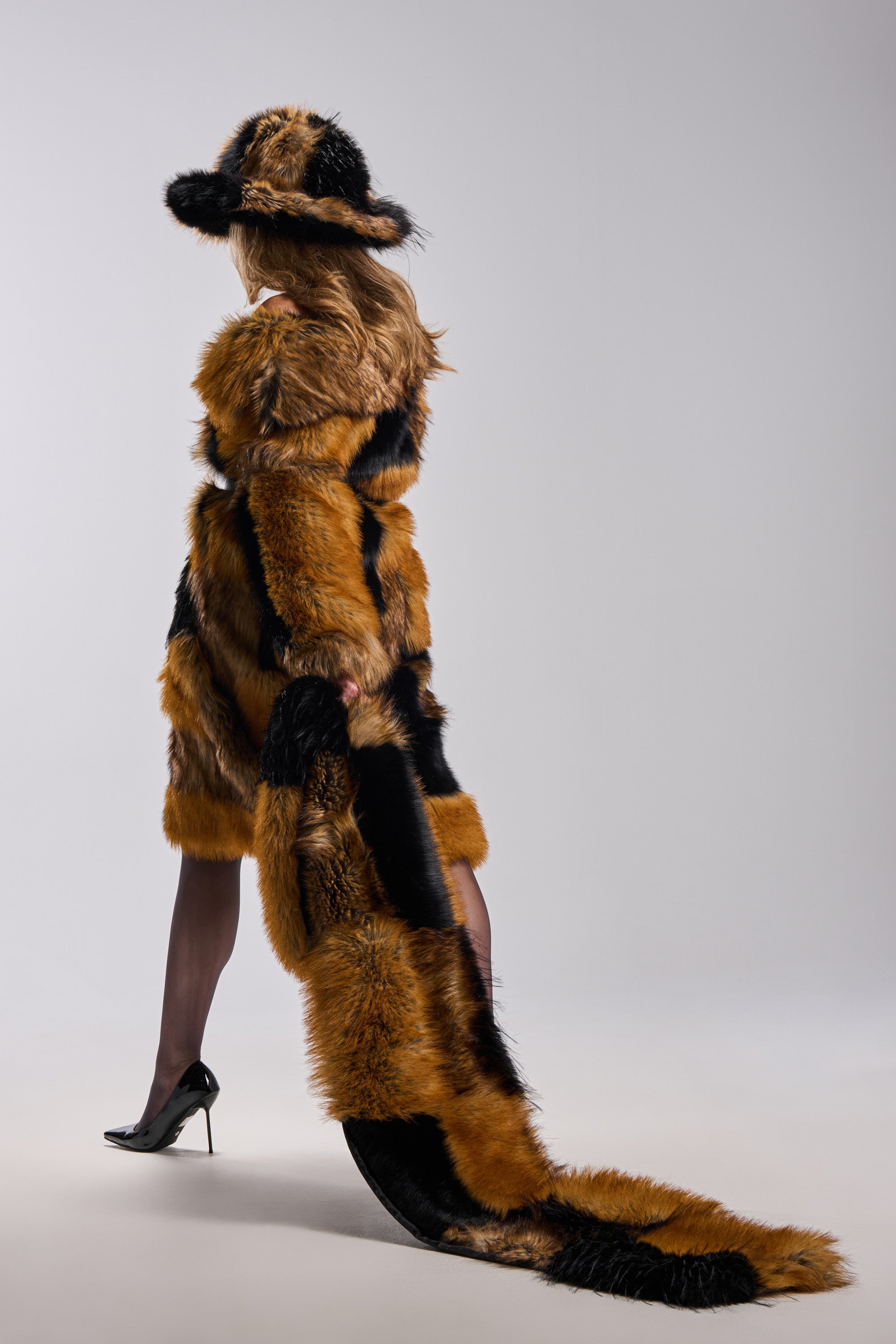A person stands against a plain light background, facing away from the camera, wearing a brown and black fur coat with a matching hat and holding the oversized COSMO FAUX FUR SCARF that trails on the floor. 1
