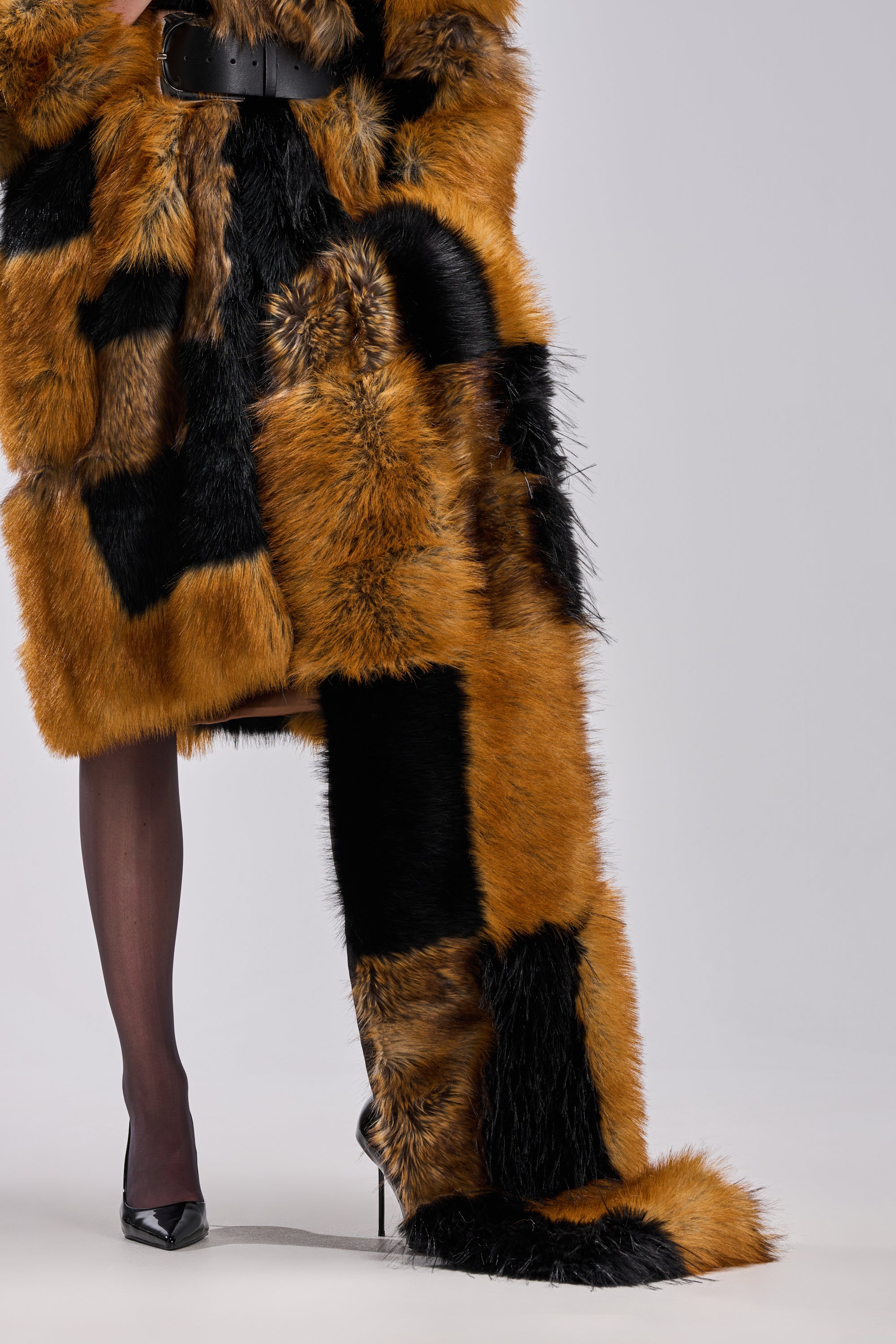 A person draped in the COSMO FAUX FUR SCARF and a black-orange patchwork fur coat with a floor-length train stands against a light gray background, revealing one leg in sheer black stockings and a black stiletto.