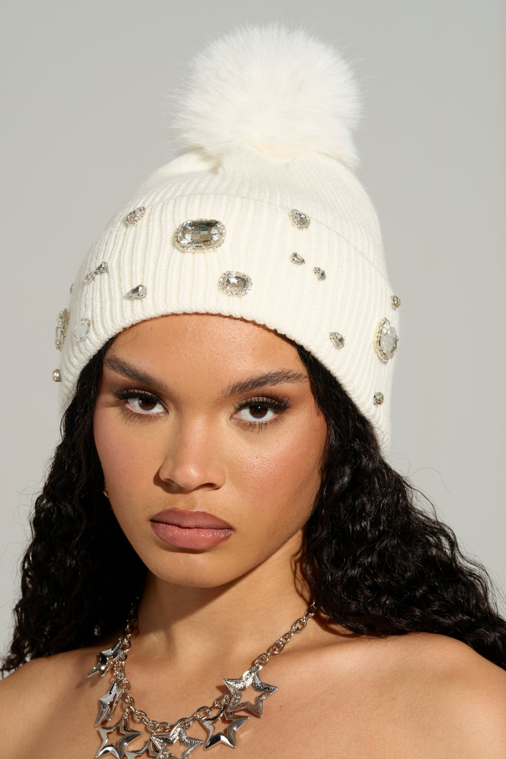 A person with long curly hair wears the Diamond Hour Embellished Beanie in white, featuring jewels and a faux fur pom-pom. They have a silver star necklace and neutral makeup, including winged eyeliner and lipstick. The background is plain and light-colored.