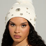 A person with long curly hair wears the Diamond Hour Embellished Beanie in white, featuring jewels and a faux fur pom-pom. They have a silver star necklace and neutral makeup, including winged eyeliner and lipstick. The background is plain and light-colored.