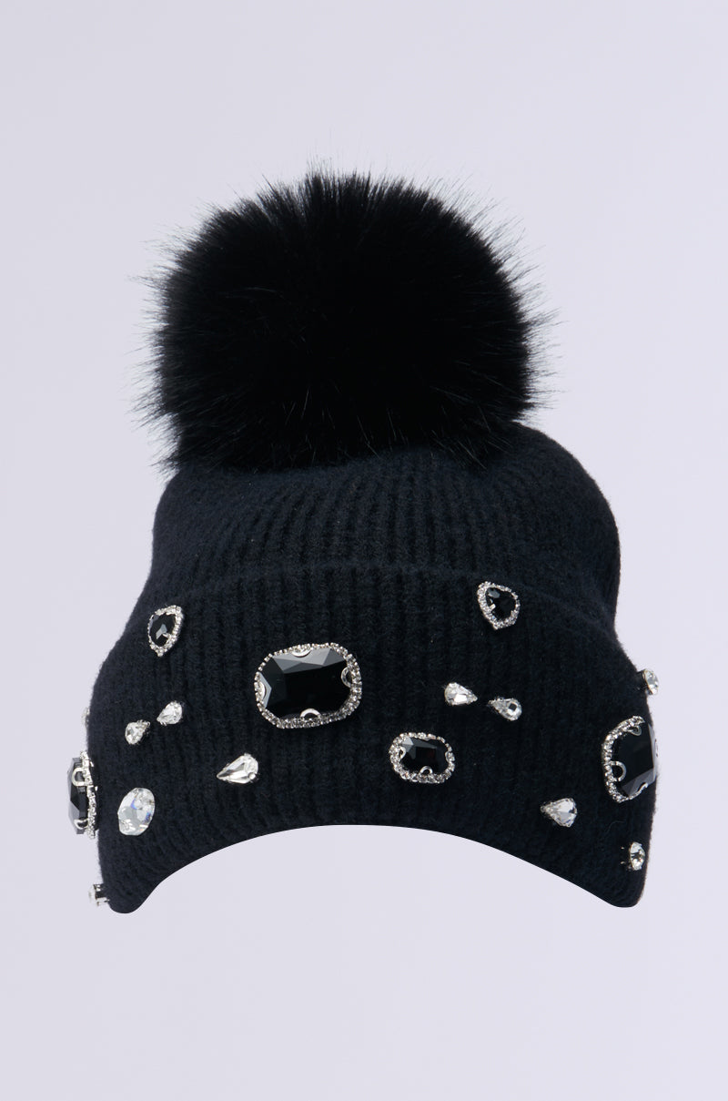 Introducing the DIAMOND HOUR EMBELLISHED BEANIE IN BLACK: a stylish knit beanie featuring a gemstone-embellished brim and an eye-catching faux fur pompom. Set against a light grey background, it's perfect for adding some sparkle to your look.