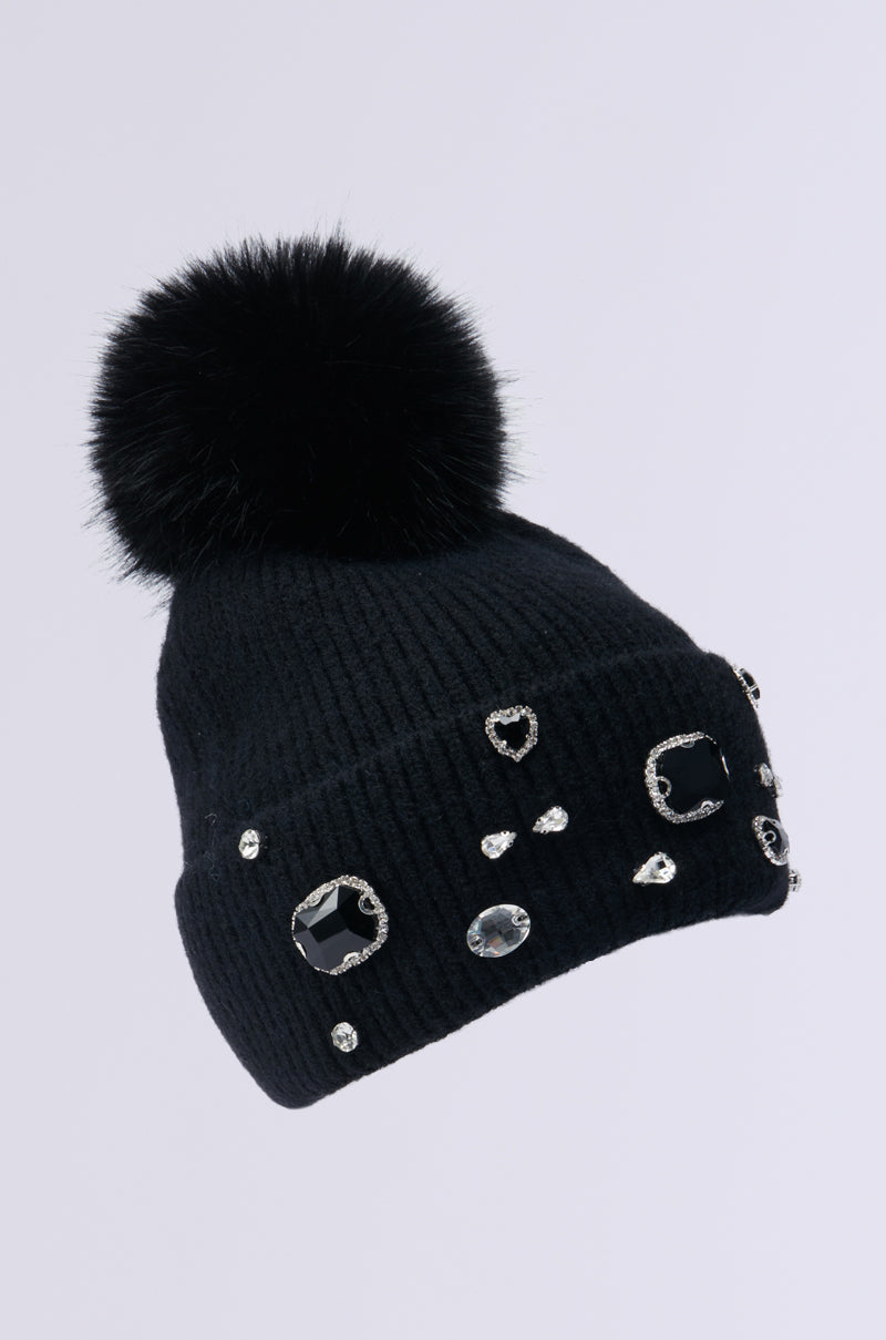 Introducing the DIAMOND HOUR EMBELLISHED BEANIE IN BLACK: a stunning knit creation in black, adorned with an intricate pattern of variously shaped rhinestones. It features a brim enriched with gemstones and is crowned with a large faux fur pompom. The rhinestones form an eye-catching design against the chic light gray background.