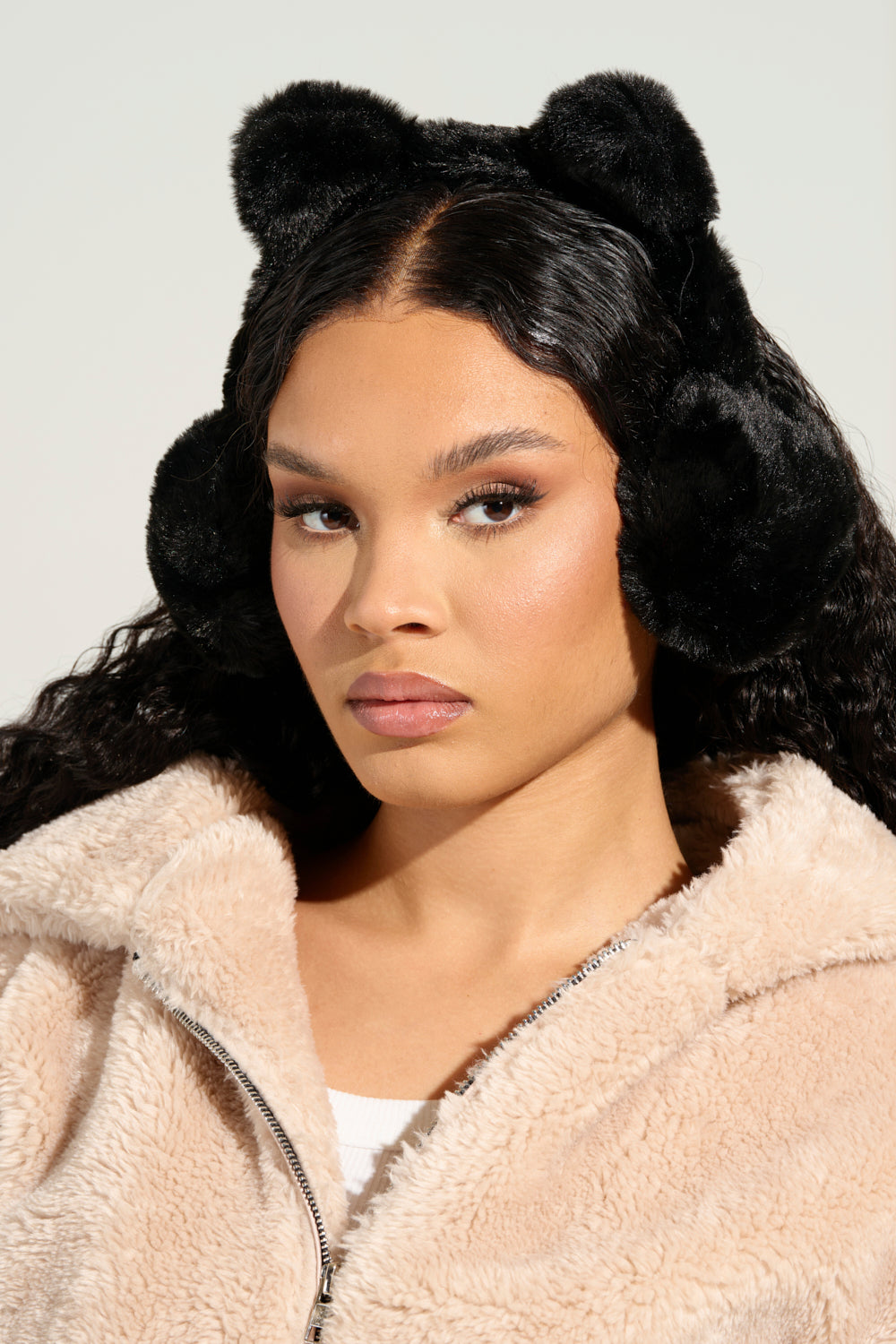 A person with long, dark curly hair wears MEOW EAR MUFFS shaped like bear ears and a beige faux fur jacket, looking at the camera against a neutral background, exuding cozy elegance.