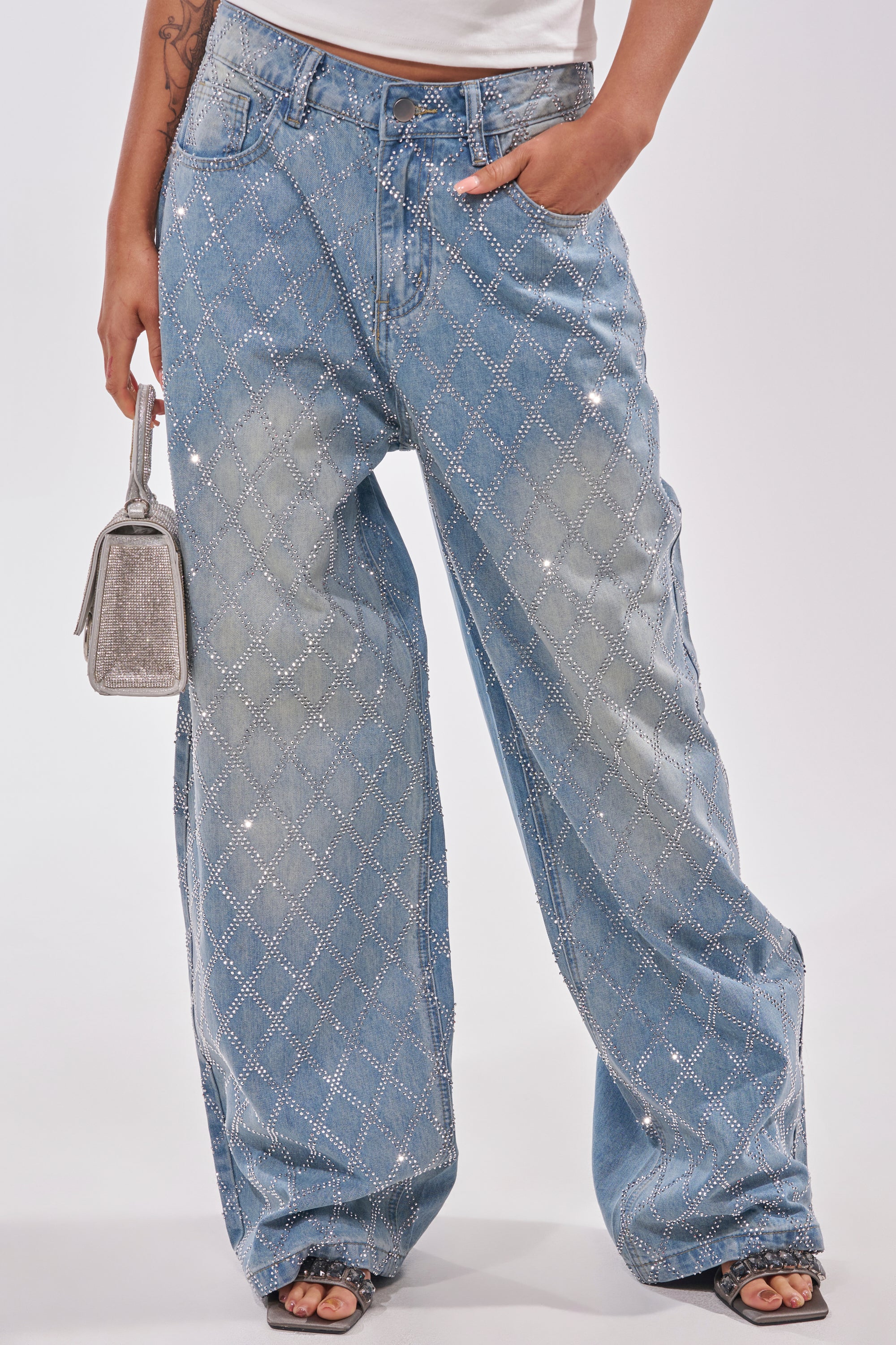 Wearing SHINE BRIGHT RHINESTONE DENIM high-rise light blue jeans with a lattice rhinestone design, a person holds a small silver handbag and stands in black open-toe shoes against a plain background. 1