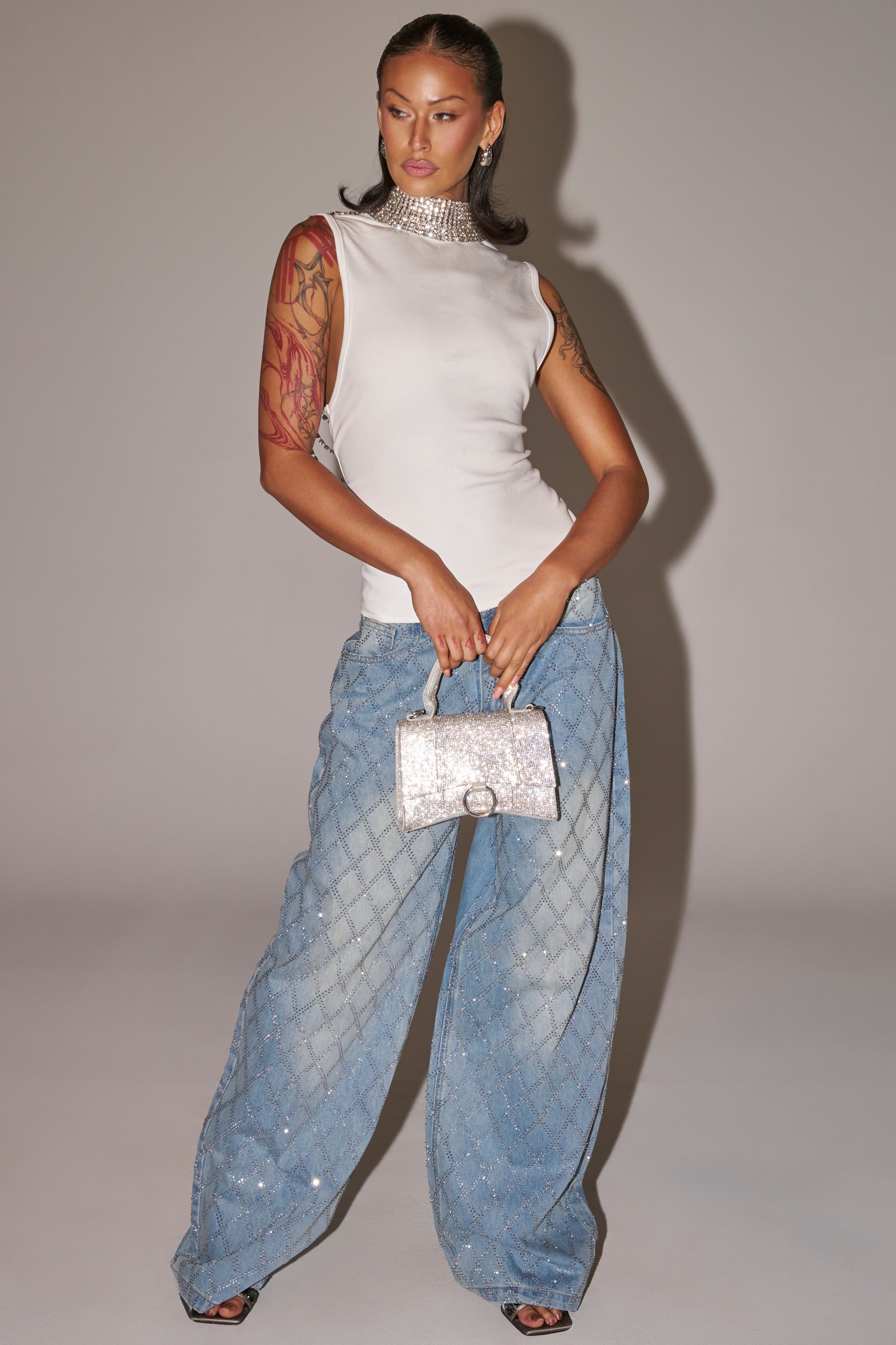 A woman poses against a neutral background wearing SHINE BRIGHT RHINESTONE DENIM, a sleeveless white top, a statement choker necklace, and holding a glittery handbag. 4