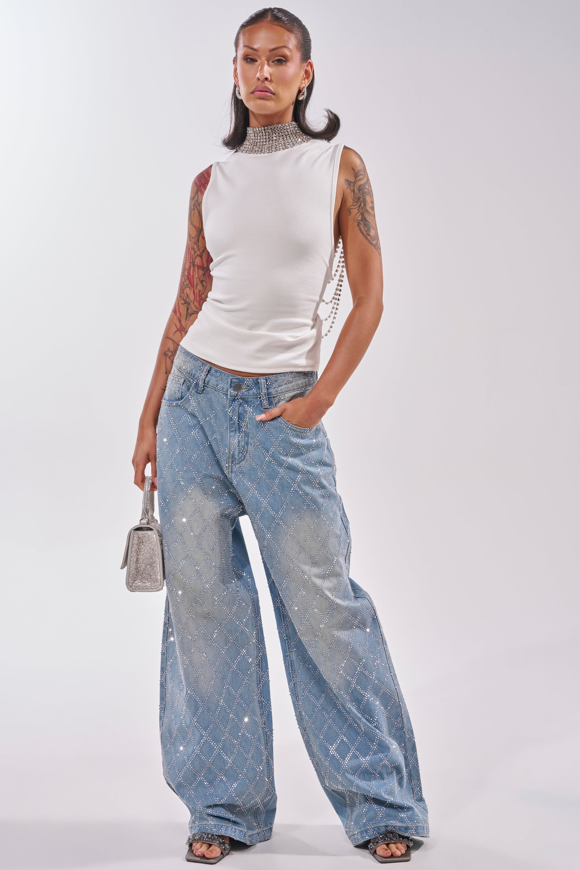 A woman stands confidently in a sleeveless white top with a high, beaded collar, SHINE BRIGHT RHINESTONE DENIM jeans, black heels, and a small silver handbag. Tattoos are visible on her arm. 5
