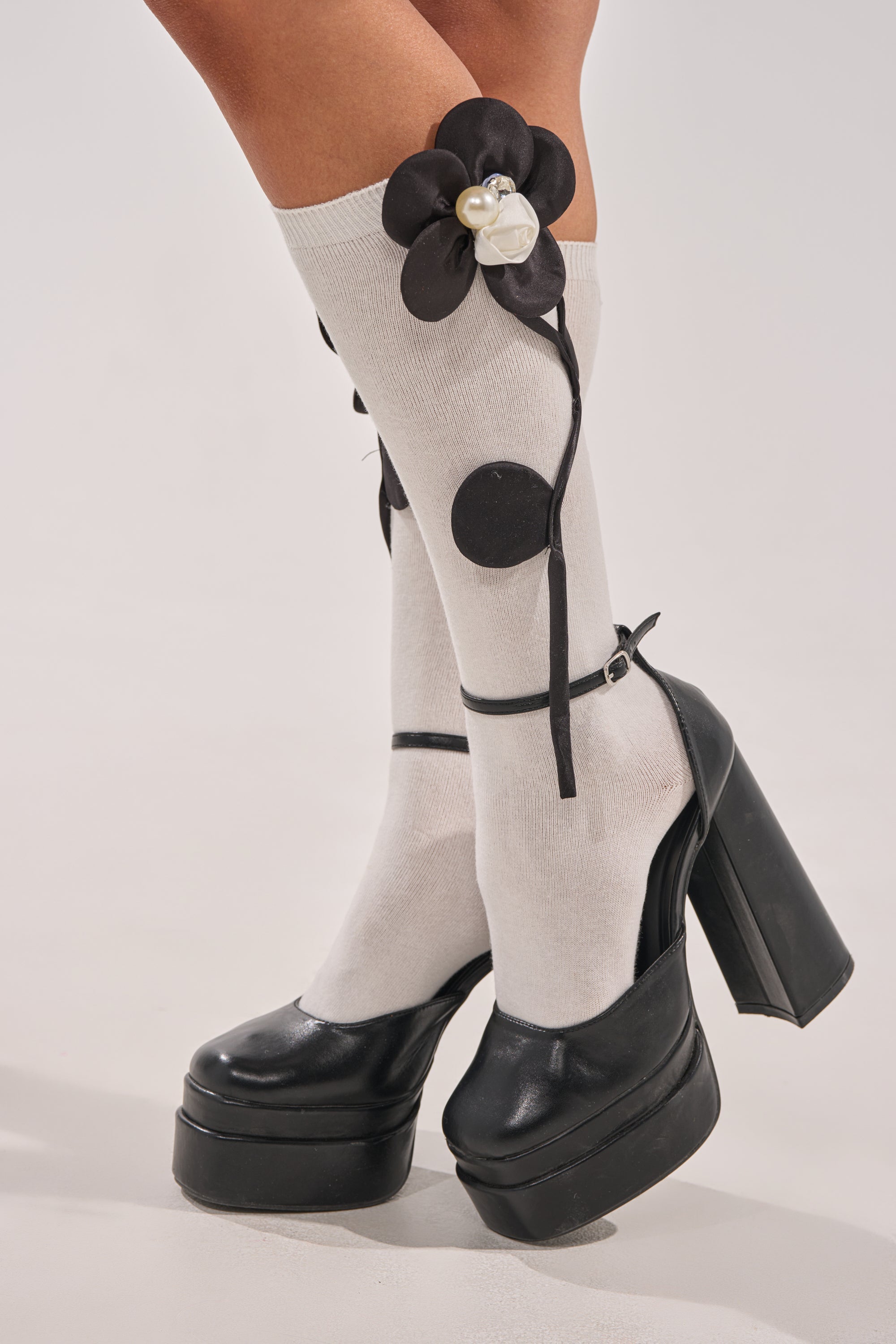 A person models the FLOWER POWER APPLIQUE SOCKS—white knee-highs with black floral appliques and rhinestone pearl accents—paired with black high-heeled platform Mary Janes, standing on a light background. 1