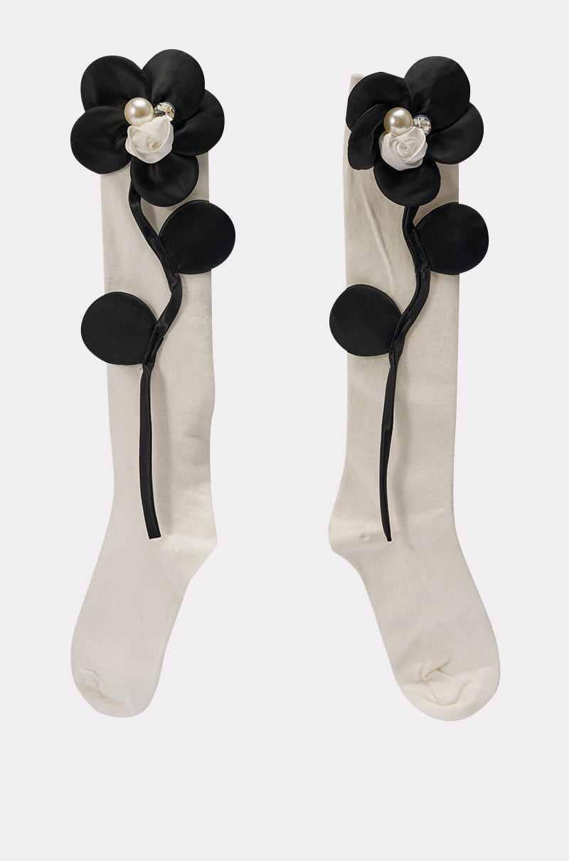 The FLOWER POWER APPLIQUE SOCKS are white, knee-high socks adorned with large black floral appliques, faux pearl and rhinestone accents, plus black circular and stem-like side embellishments. 2