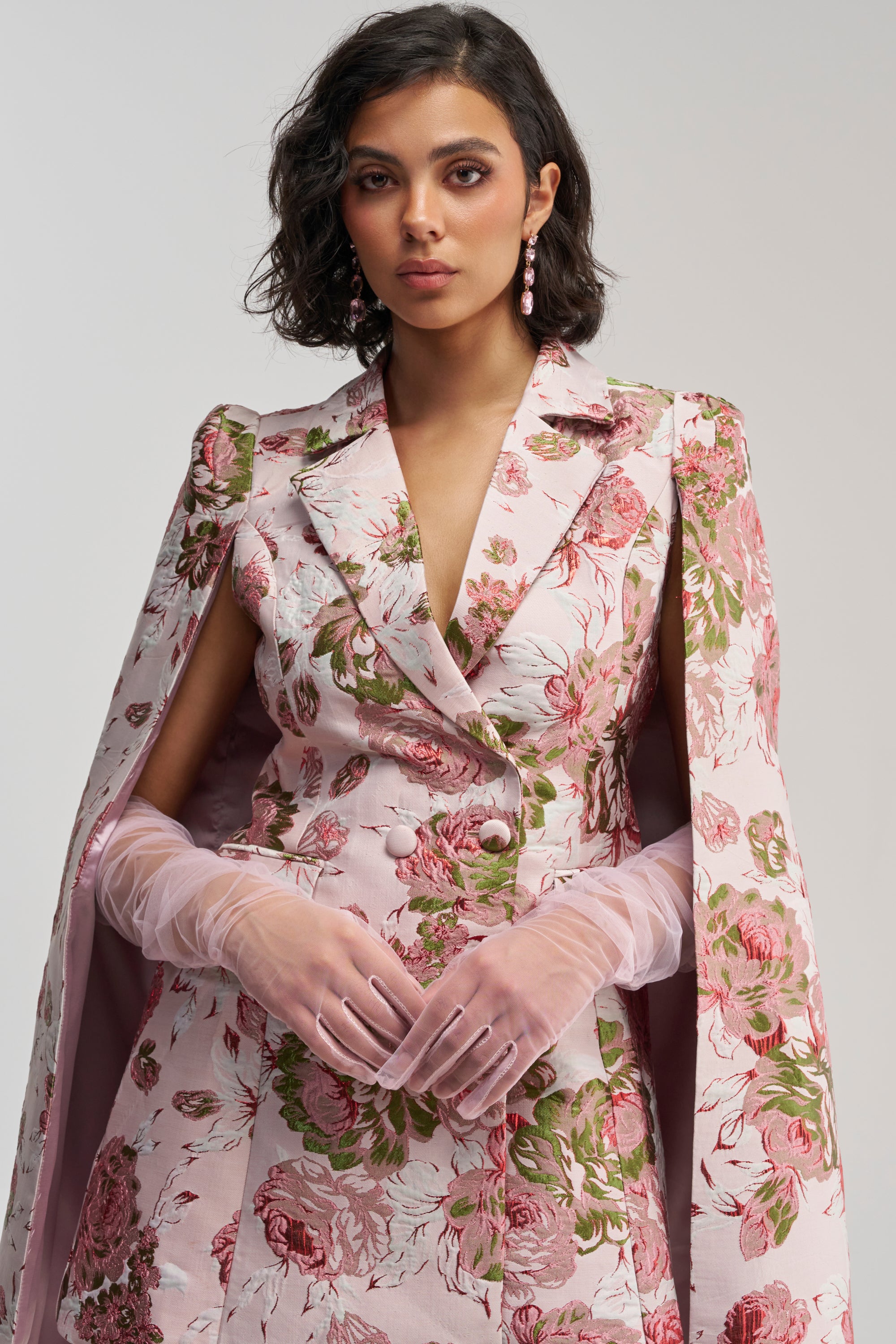 A woman with short dark hair wears a pink floral suit with a matching cape. She accessorizes with the SHEER EMOTION MESH GLOVES (PINK), elegant drop earrings, and stands confidently against a plain light background.