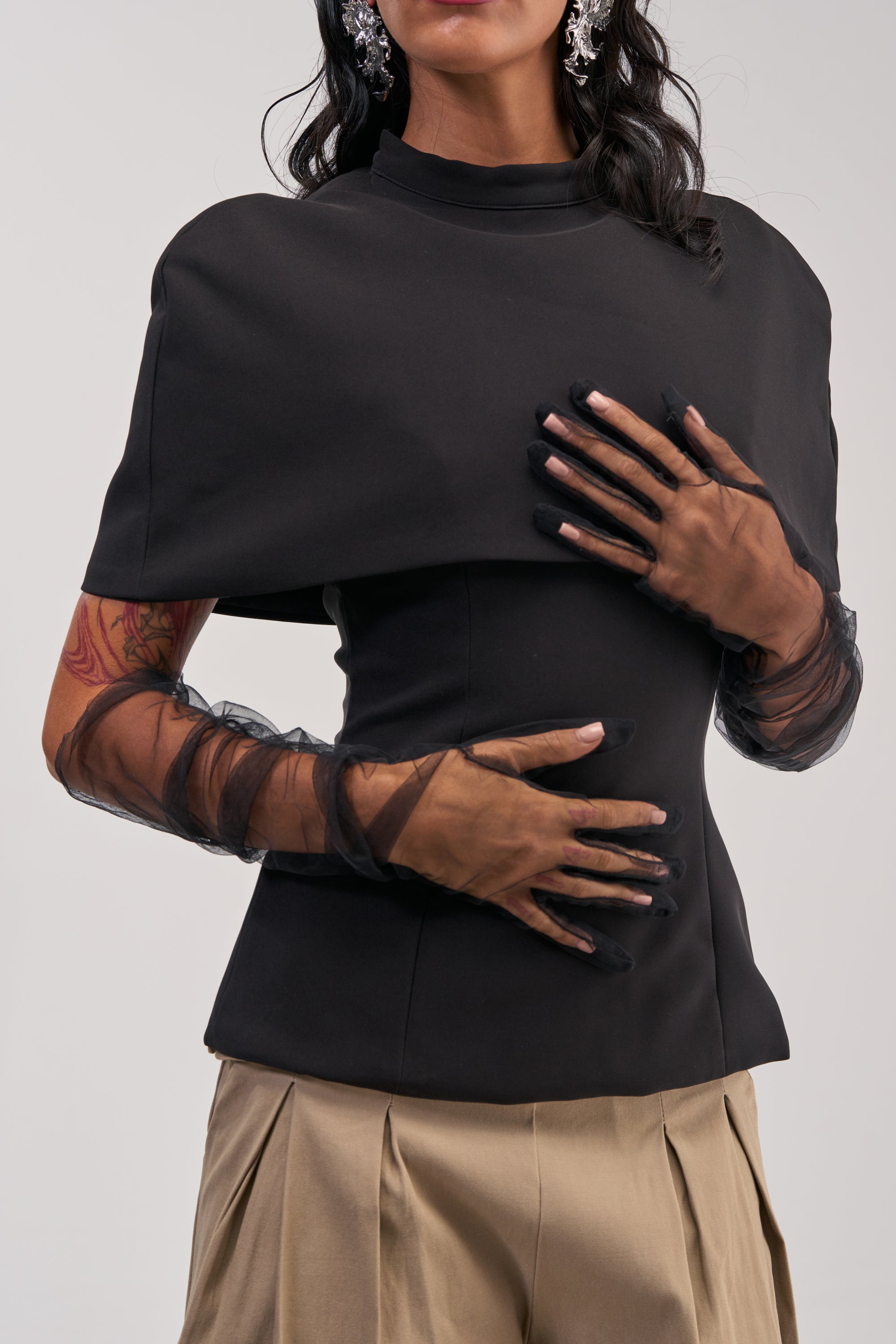 A woman wears the SHEER EMOTION MESH GLOVES IN BLACK with a black top, beige pleated pants, and statement earrings. Her visible arm tattoos stand out as she holds her torso with both hands against a plain, light background.