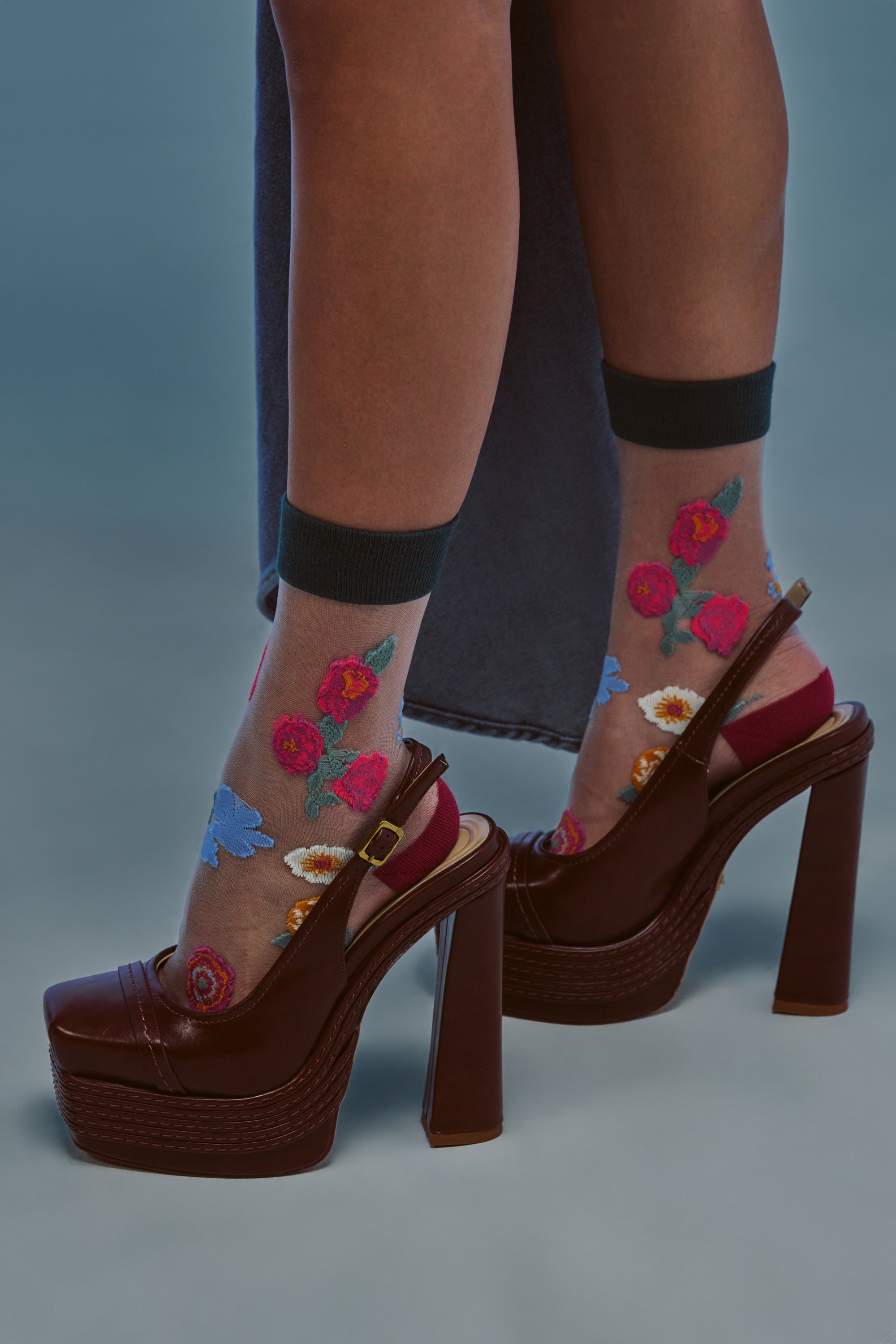 A person wearing SO DELICATE SOCKS with colorful floral embroidery and maroon platform high heel sandals stands on a grayish-blue surface.