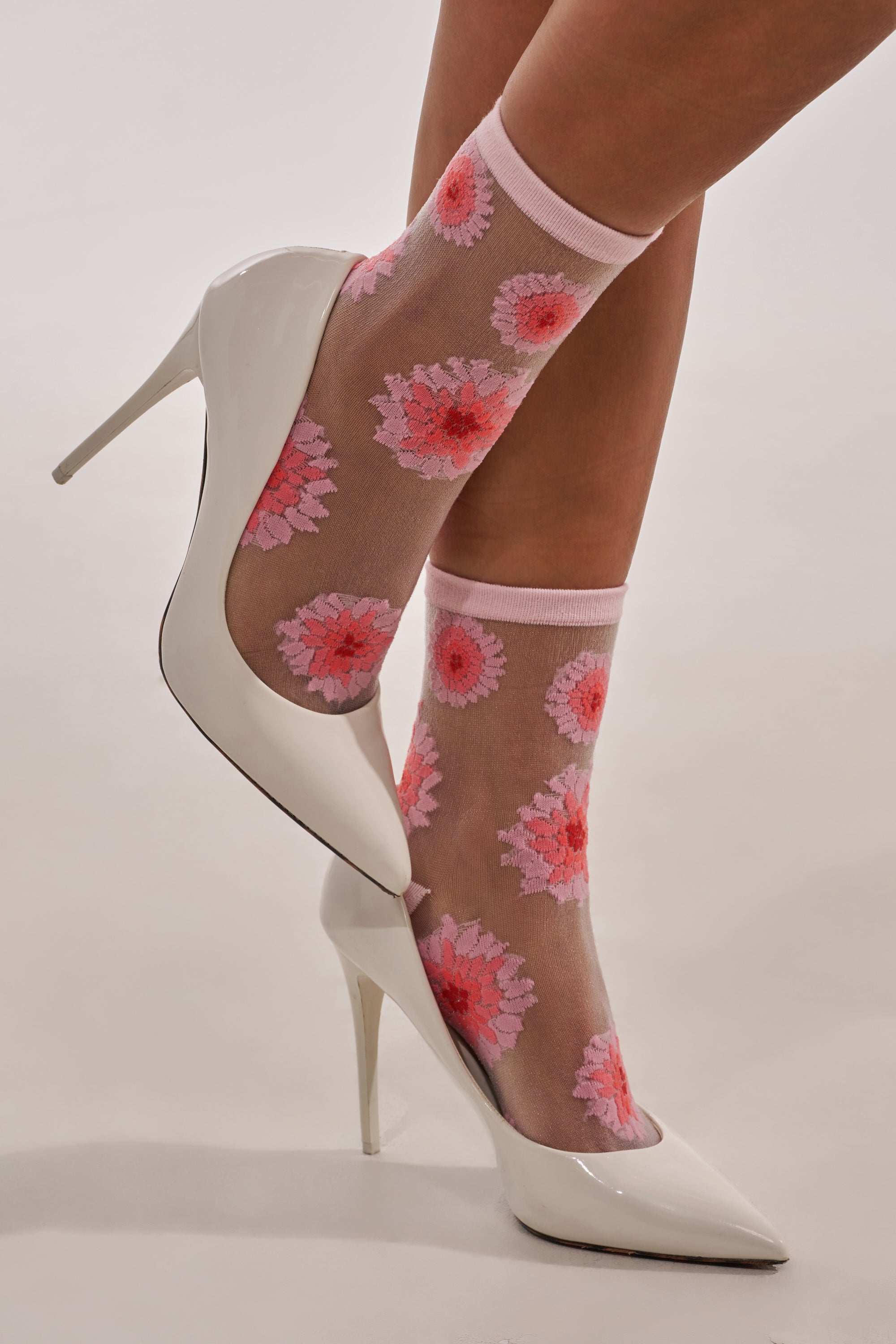 Someone stands against a plain light background wearing WHERES MY HEEL SOCKS, featuring a cute mesh floral pattern with pink and red flowers, paired with white high-heeled shoes. 0